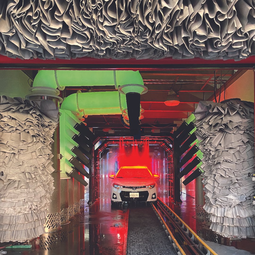 A white car is in an automated car wash station with red and green colored lighting, and spinning brushes with ruffled fabric coverings.