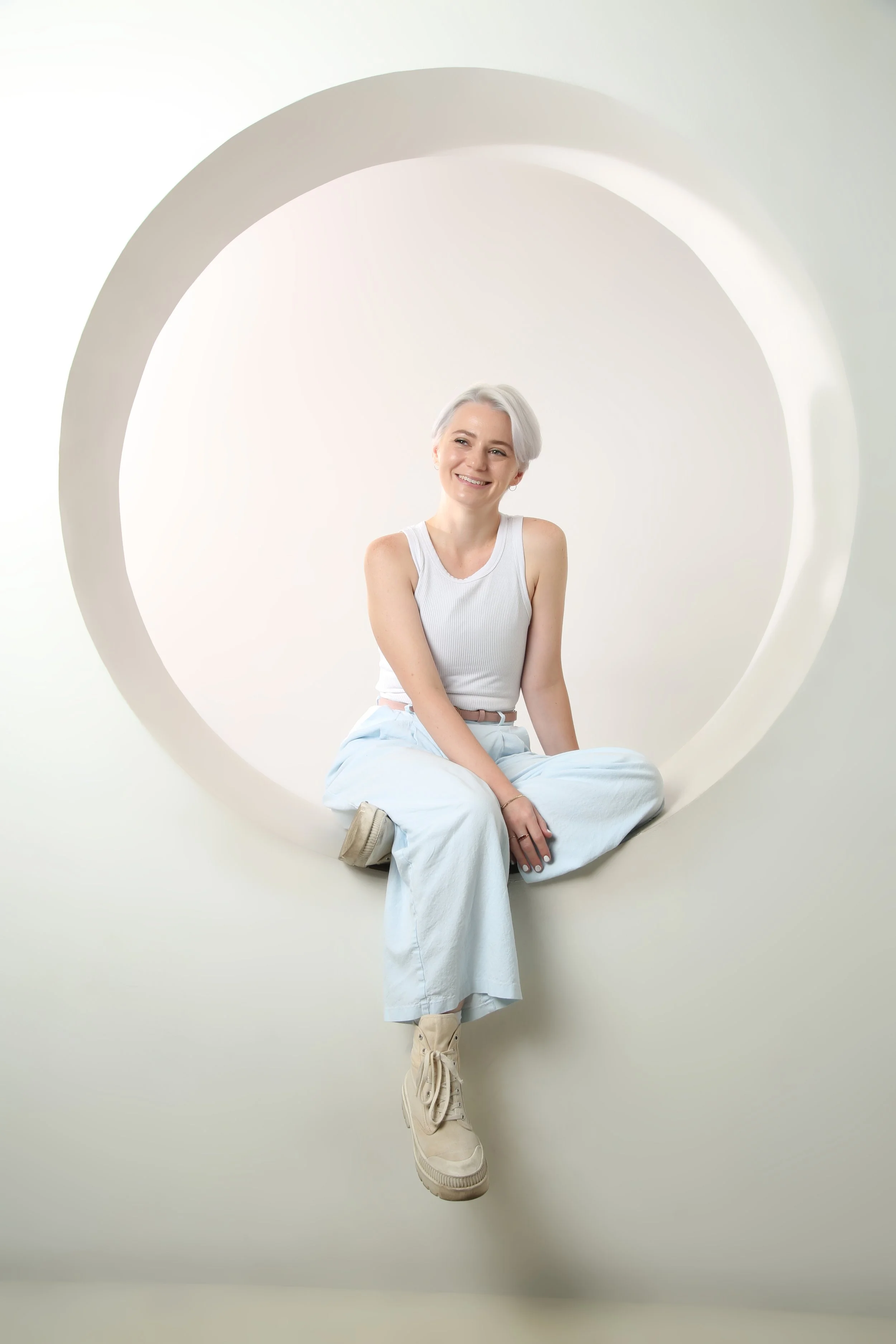 A trans man with short grey hair wearing a white tank top and light blue pants, sitting inside a white circular tunnel, smiling.