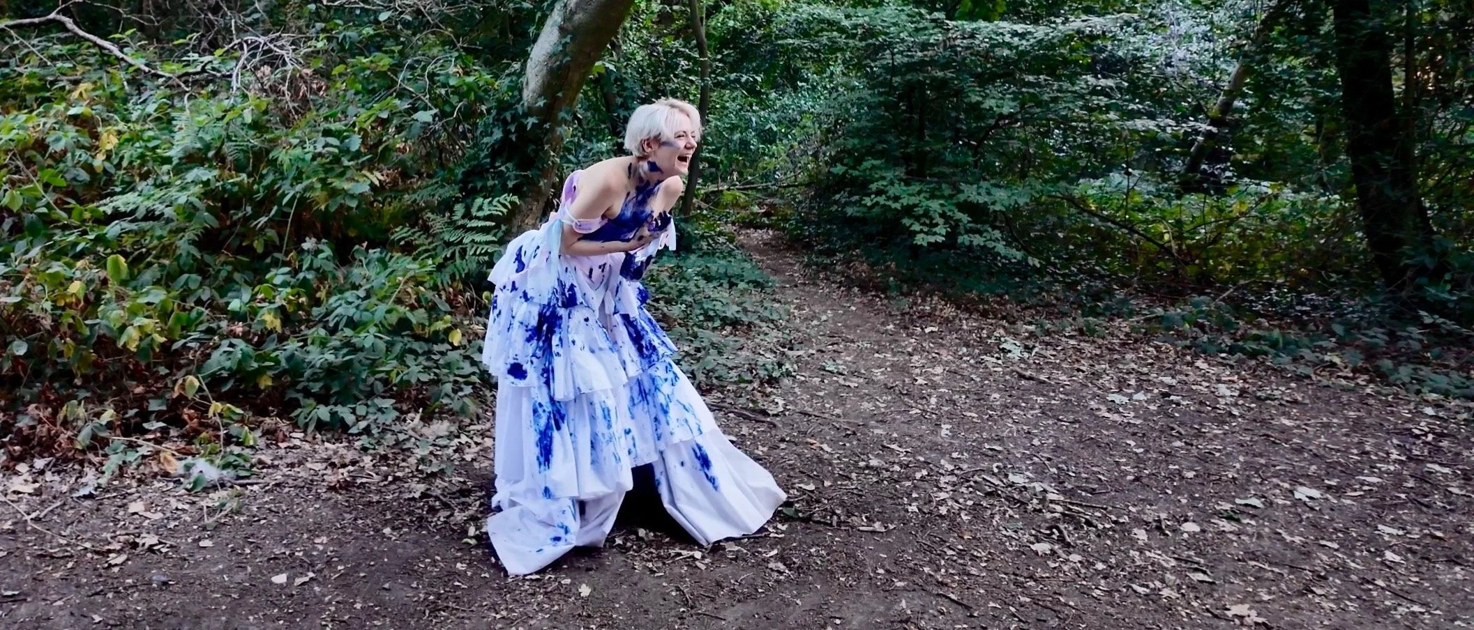 A trans man in a white and blue dress stands on a forest trail, clutching his stomach and laughing.
