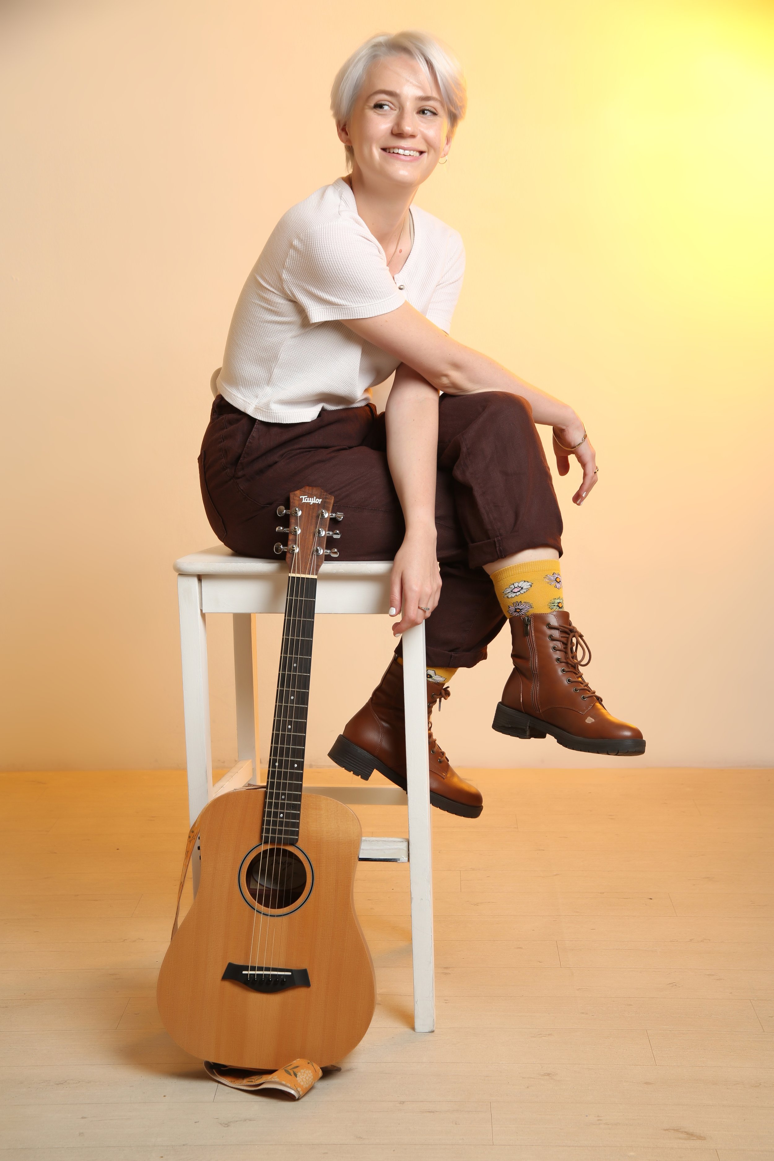 short gray hair sitting on a white stool, smiling, wearing a white t-shirt, dark jeans, and brown lace-up boots, a guitar on the ground leaning against the stool, and yellow socks with flowers. The background is a plain white wall with warm lighting.