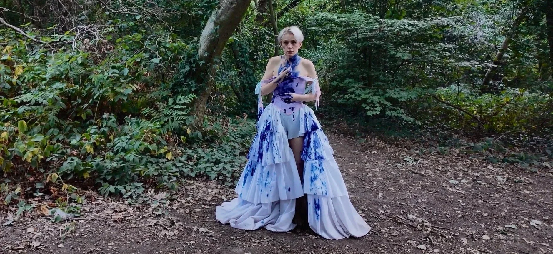 A trans man with short platinum blonde hair standing on a forest trail, wearing a strapless dress with layered white fabric painted with blue splatters.