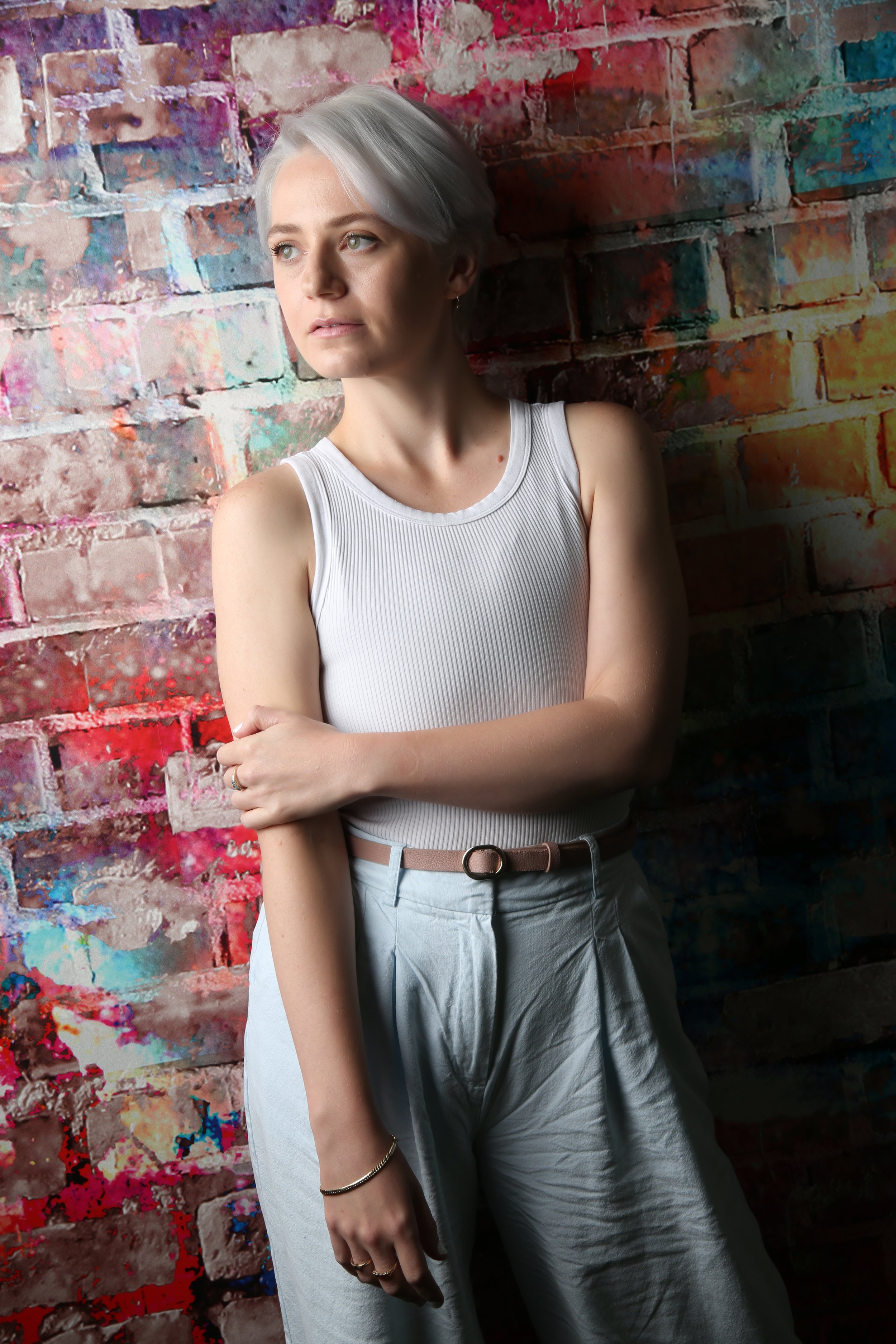 A trans man with short, light grey hair and wearing a white sleeveless top and light grey pants with a brown belt, stands with arms crossed against a colourful, painted brick wall.
