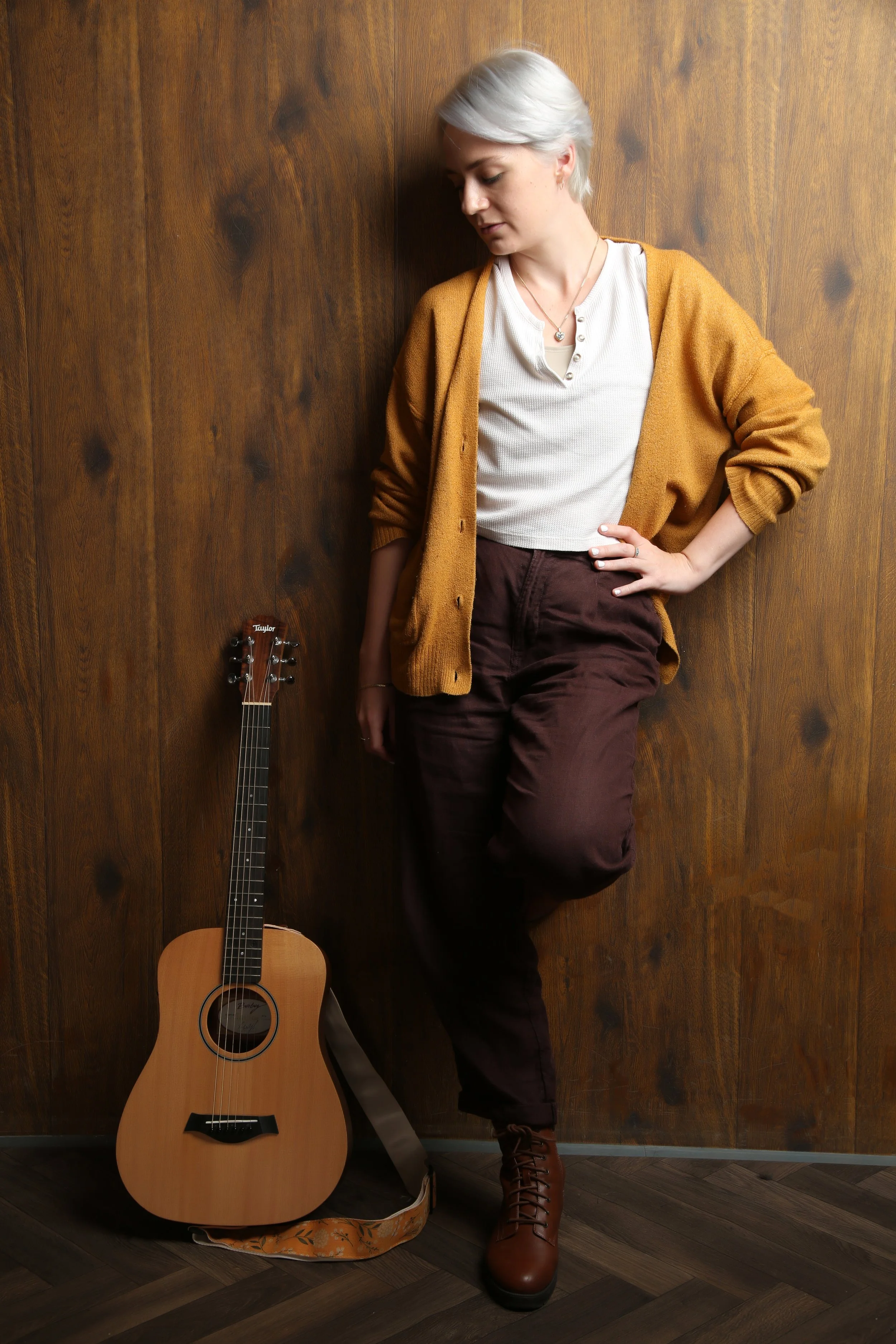 A trans man with short white hair, wearing a brown cardigan, white top, and brown pants, standing against a wooden wall next to an acoustic guitar.