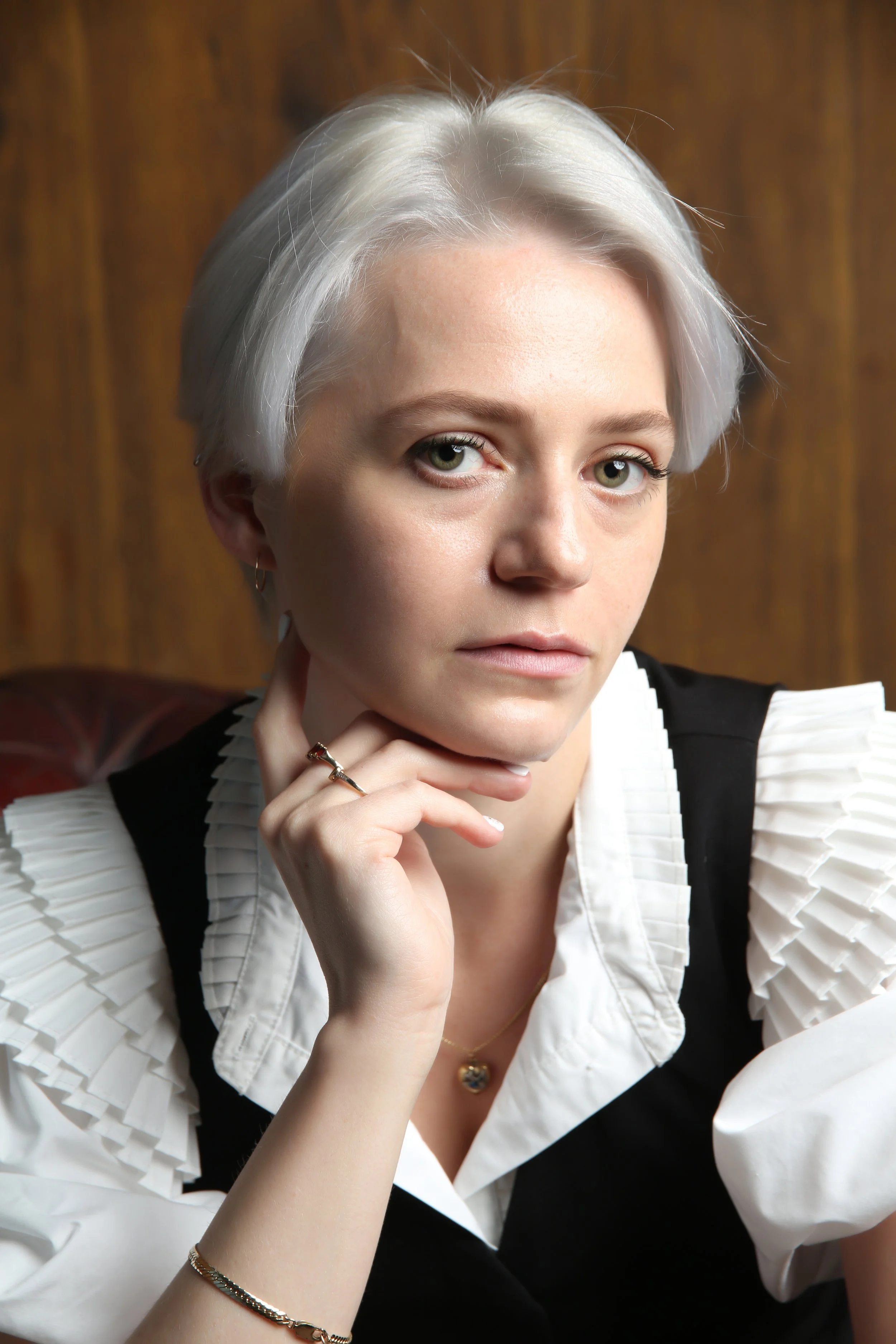 A woman with short, platinum blonde hair, wearing a white blouse with pleated sleeves and a black vest, sitting in front of a wooden background, resting her chin on her hand, wearing rings and jewelry.