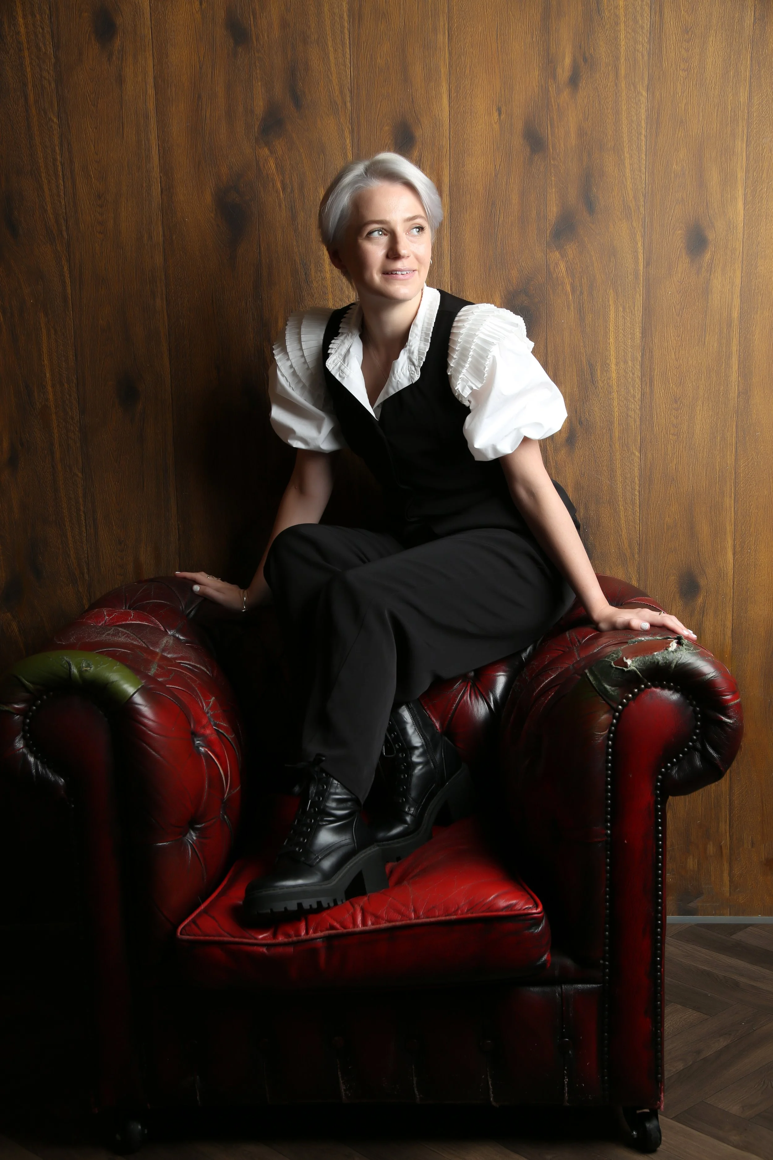 A woman with short gray hair and fair skin sitting on a vintage red leather tufted couch with a wooden wall background. She wears a white blouse with puffed sleeves and a black vest and pants, with black combat boots.
