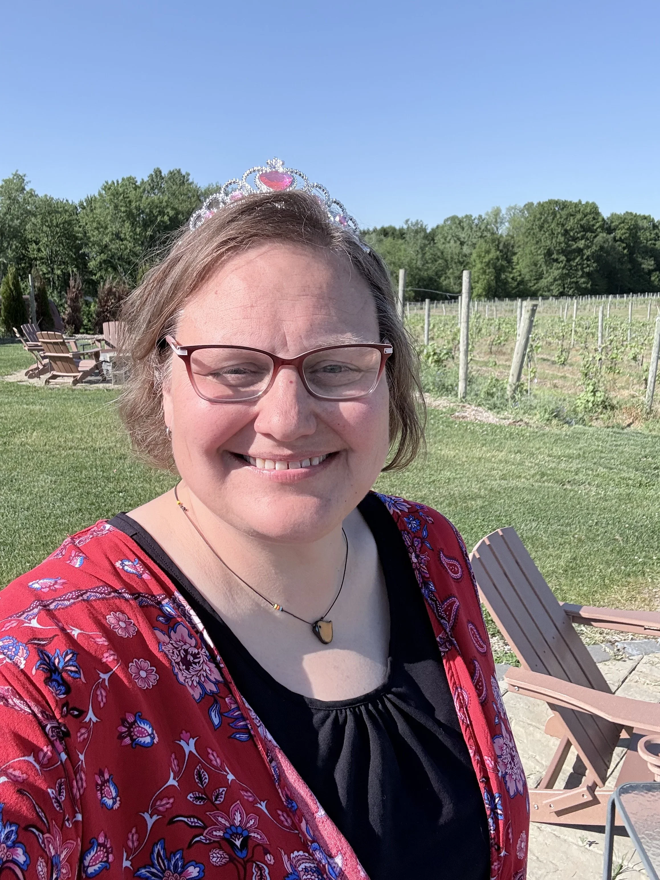 A woman smiling outdoors, wearing red glasses, a tiara, a black top, and a red floral-patterned jacket. Behind her are green trees, a blue sky, and outdoor chairs.