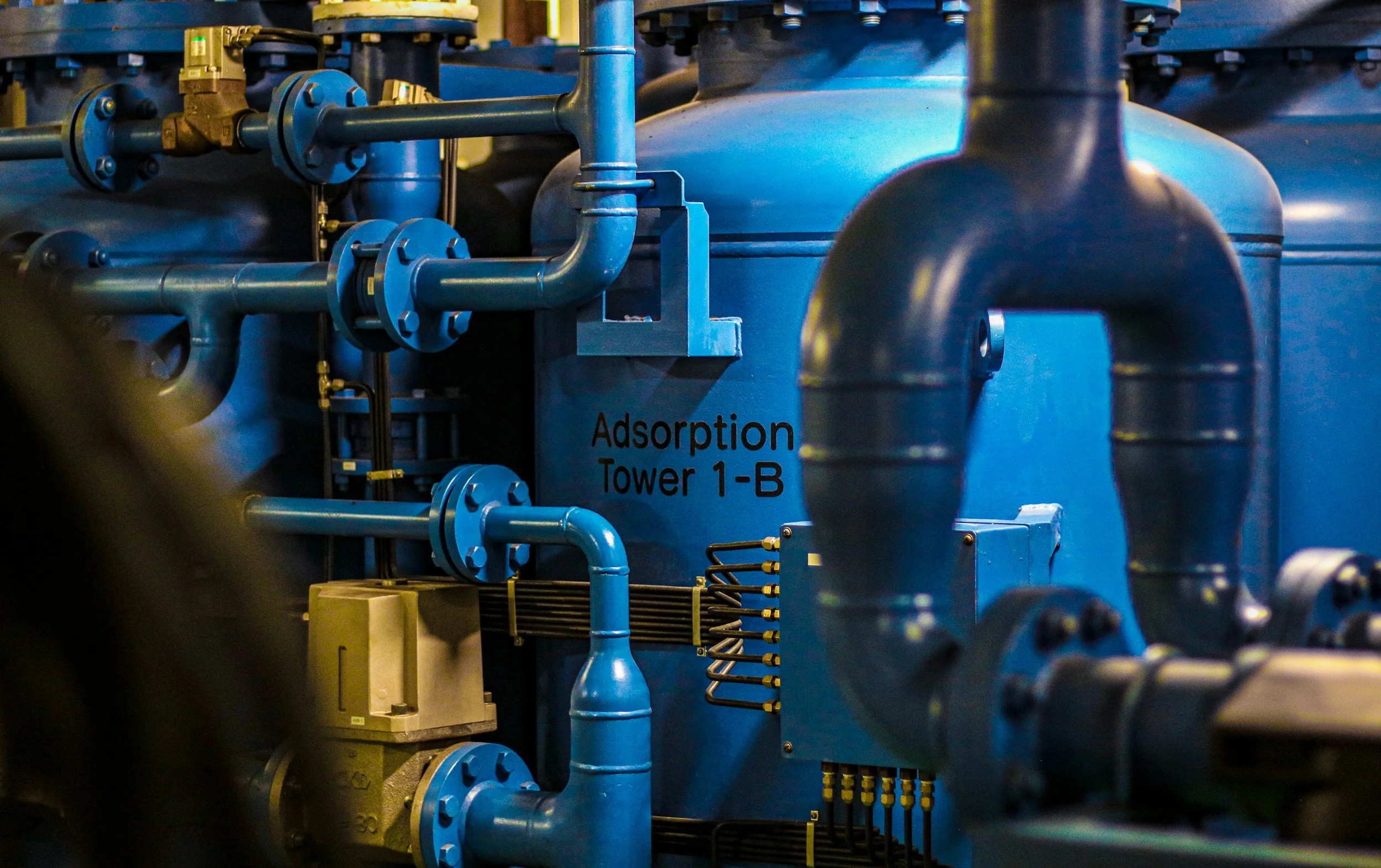 Close-up of industrial piping and machinery, including blue pipes and valves, with a label reading 'Adsorption Tower 1-B'.