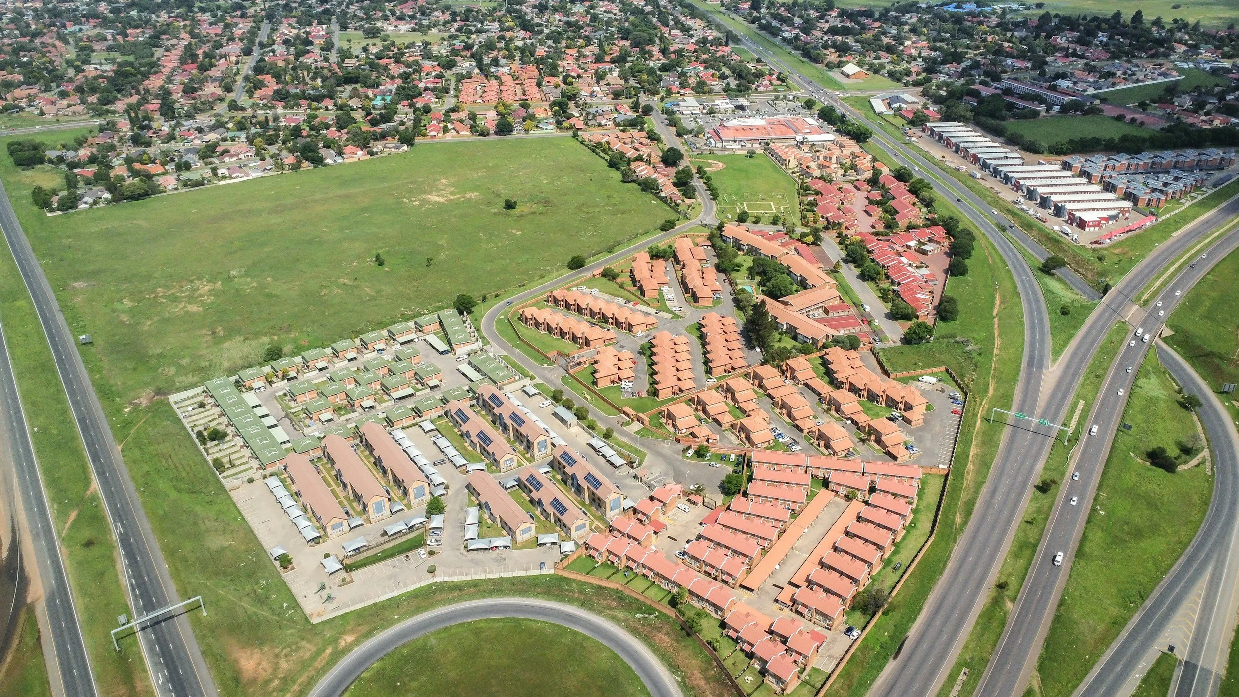 Aerial view of a suburban area with houses, green fields, and roads, including a highway with vehicles on it.