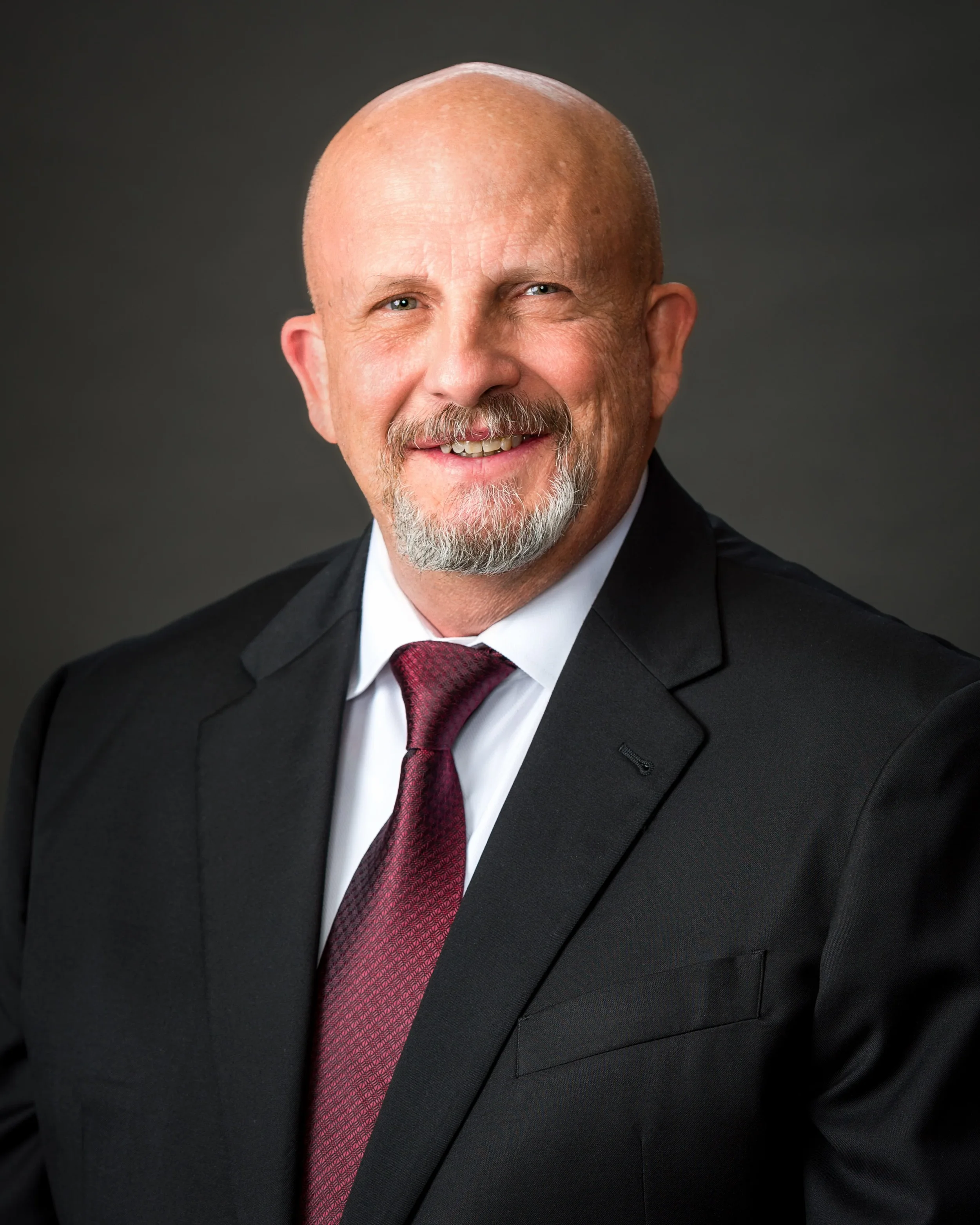 Headshot of a middle-aged man with a bald head, goatee, and mustache, wearing a black suit, white shirt, and a red tie, smiling against a dark background.