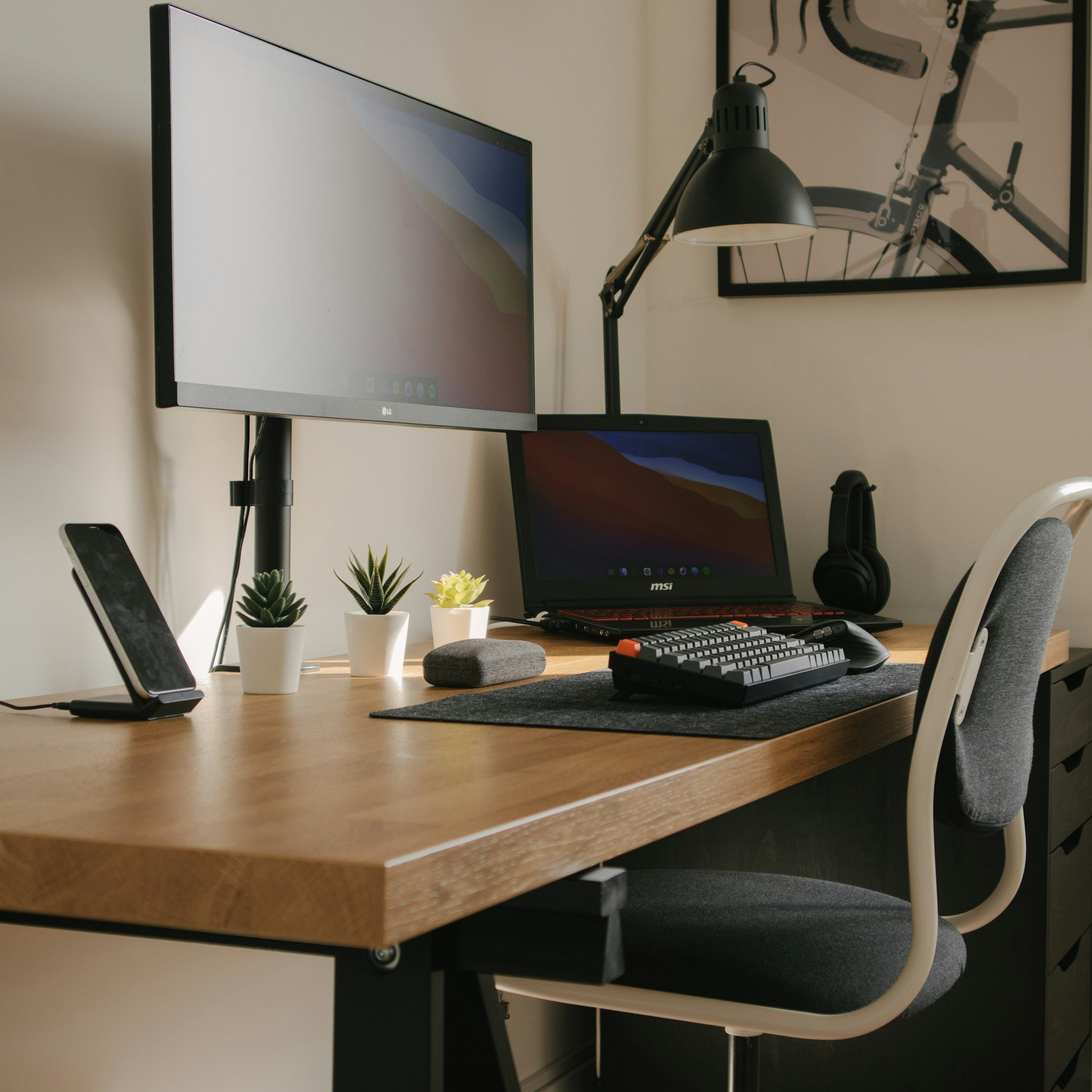 A workspace with a computer monitor, a laptop, a keyboard, a mouse, a lamp, and small potted plants on a wooden desk. There is a framed bicycle picture on the wall.