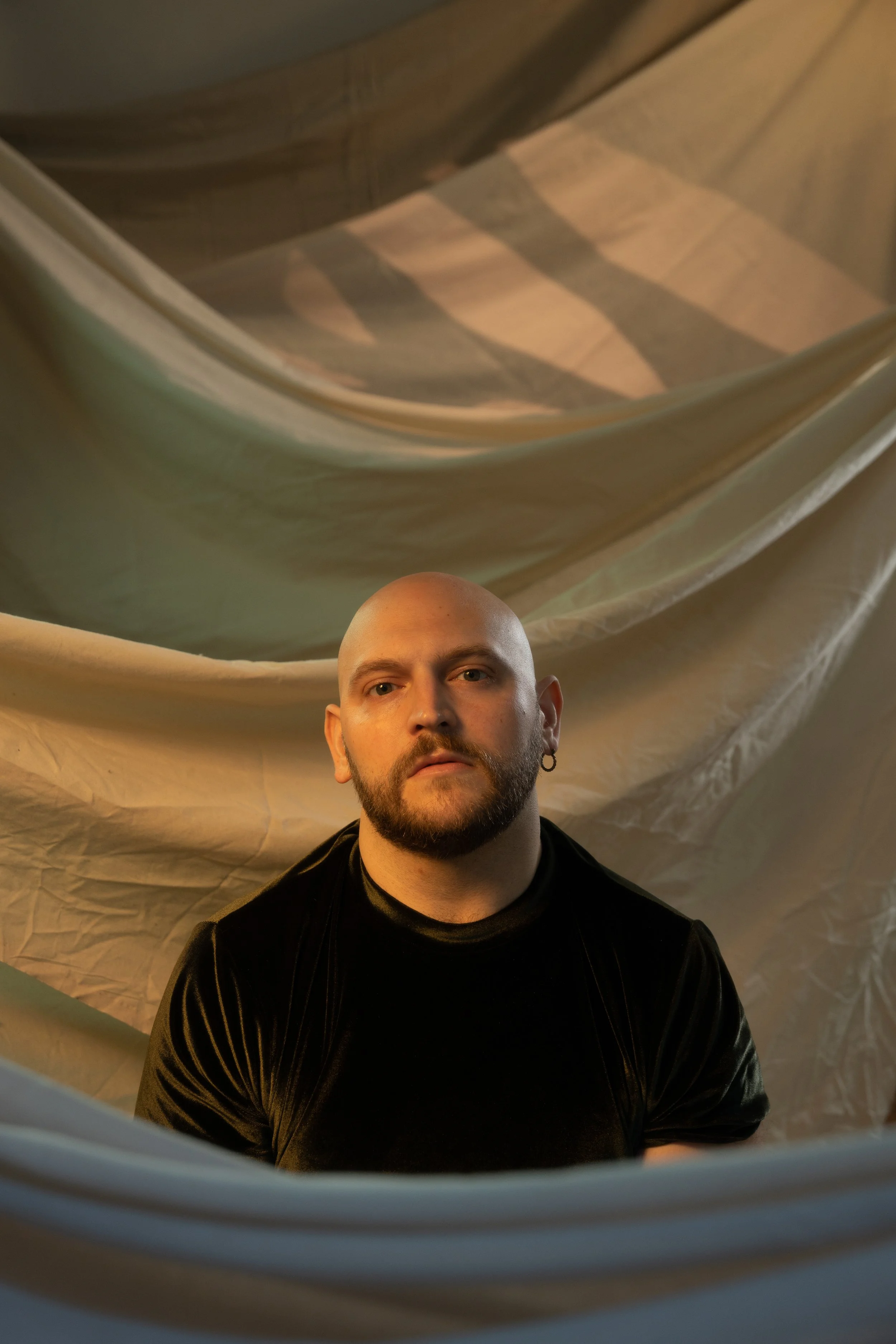 Stephan sits in a dark green velour shirt in front of a billowy tan curtain backdrop