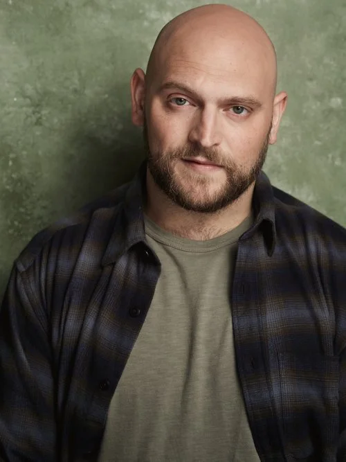 Stephan is in a blue and green flannel over a green shirt in front of a speckled green backdrop