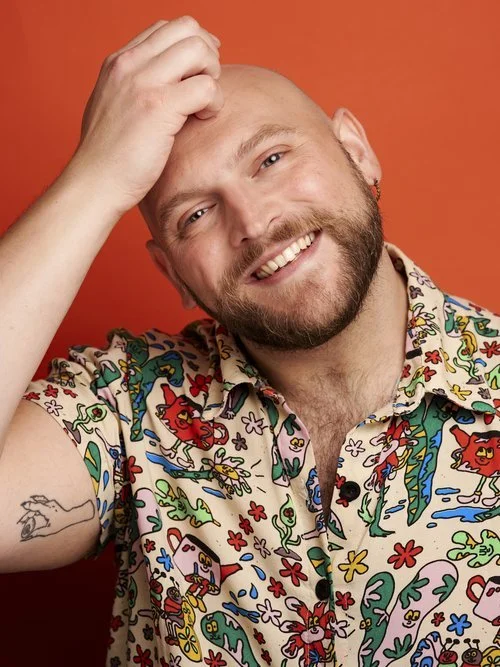 Stephan is a tan button down shirt with colorful bugs and pitchers on it in front of an orange backdrop