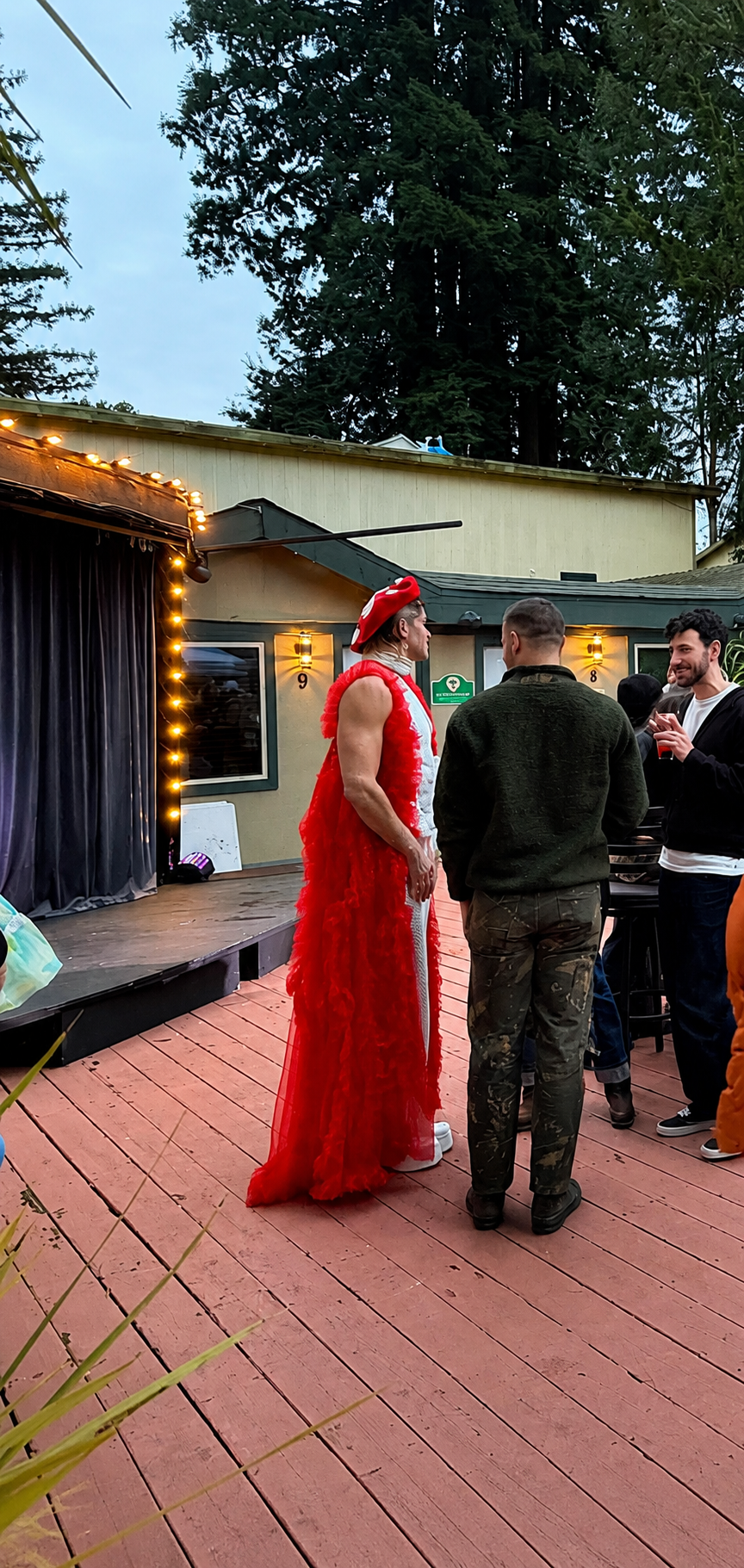 A drag queen dressed in a red dress is talking to others on a wooden deck outdoor stage area with string lights and a beige building in the background.