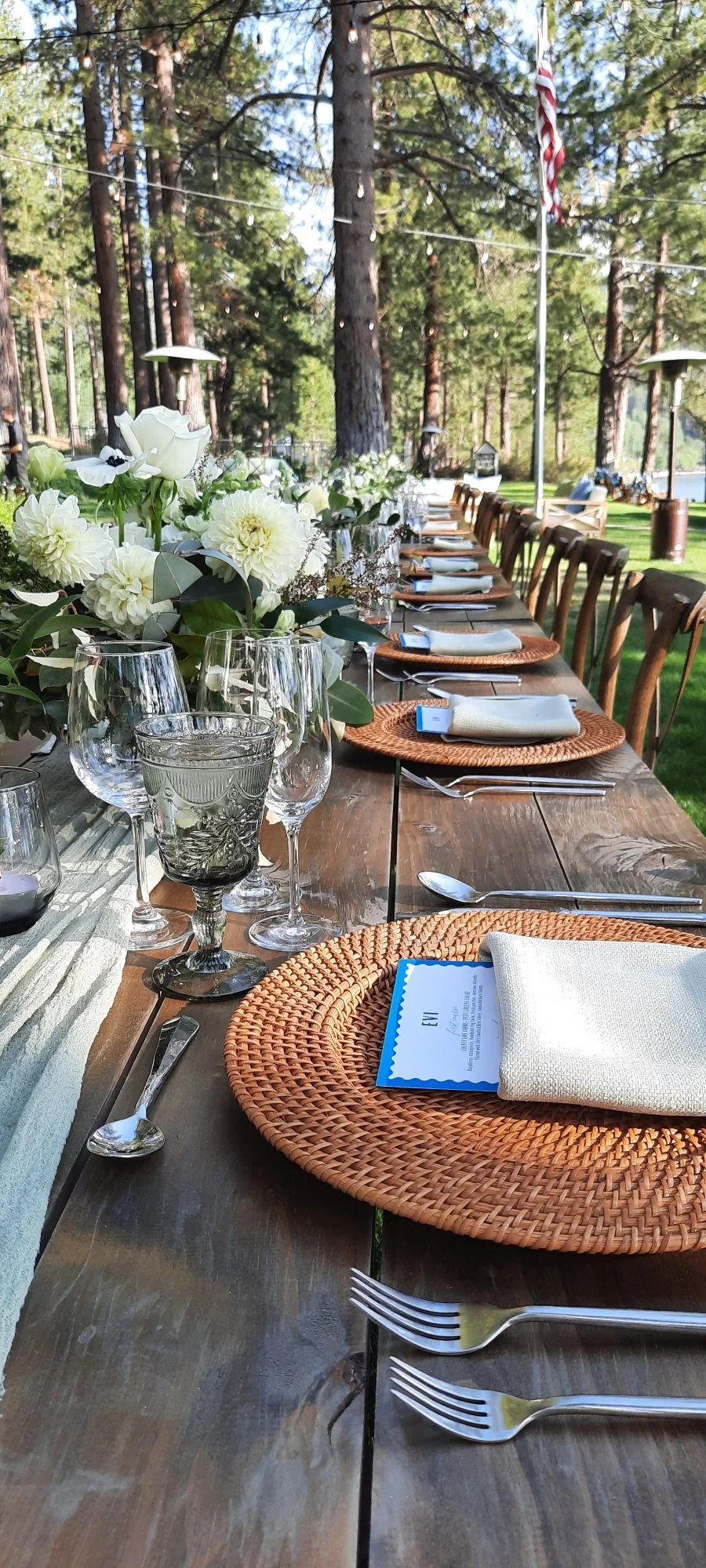 Outdoor dining setup with a long wooden table decorated with white floral centerpieces, surrounded by wooden chairs, set with woven placemats, napkins, silverware, and glassware, in a forested area with tall trees and string lights.