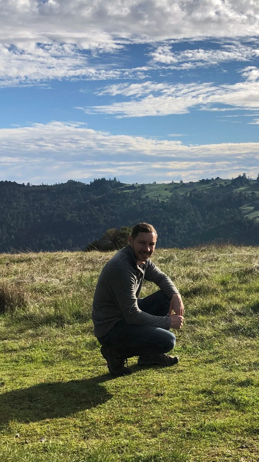 A man squatting in a grassy field with mountains and a partly cloudy sky in the background.