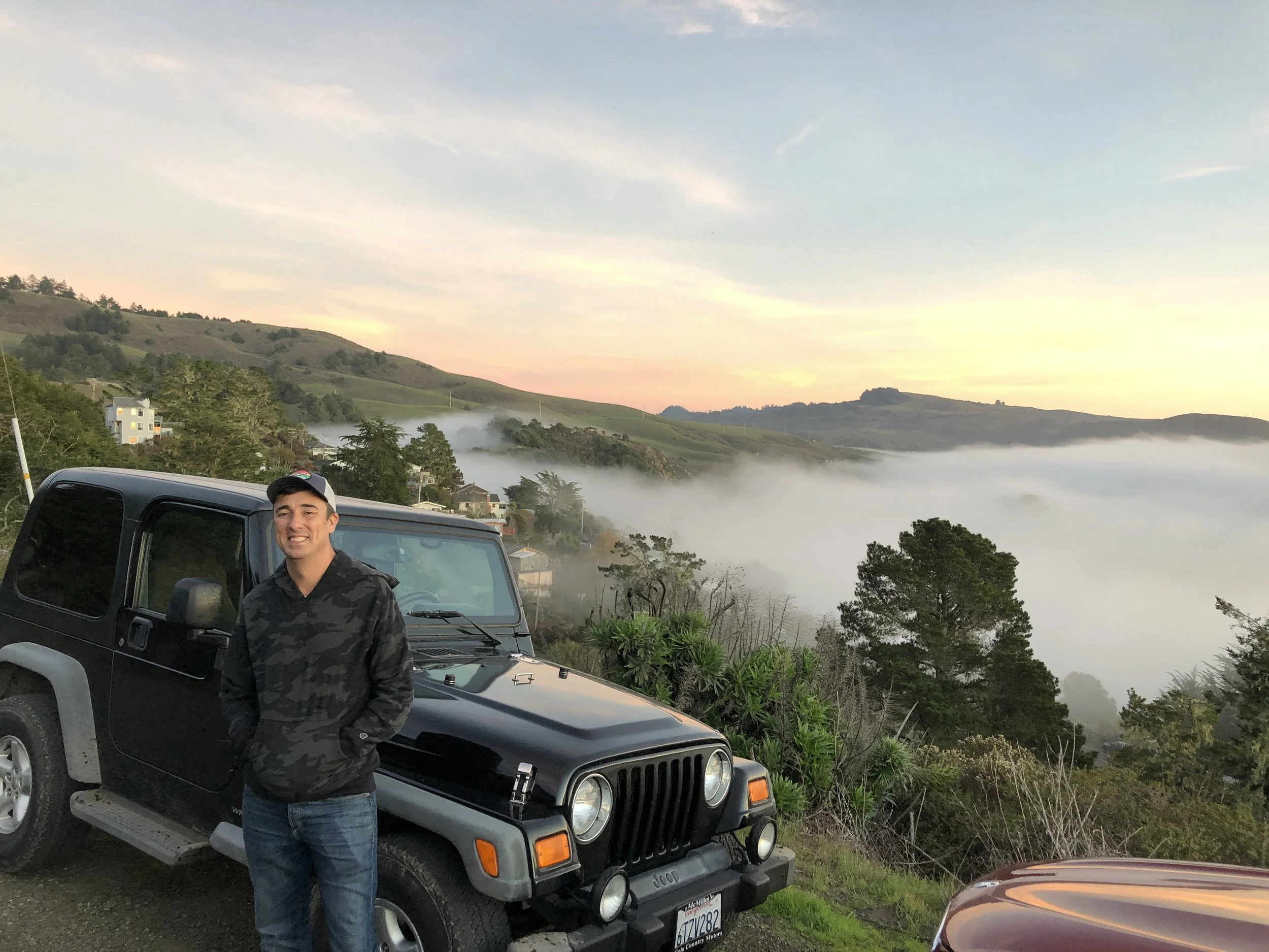 A man is standing in front of a black Jeep vehicle, smiling at the camera. Behind him, there is a hillside with houses and trees, and a layer of mist or fog near the ground. The sky above shows early morning or late evening light with soft, pastel-colored clouds.
