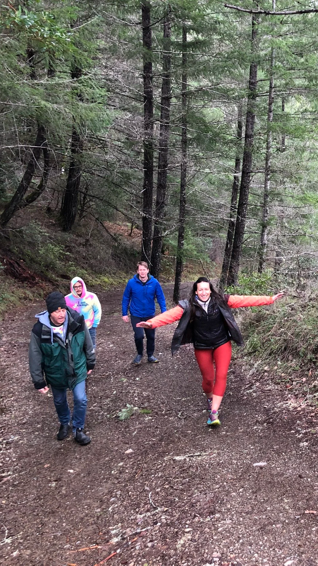 Four people walking on a redwoods trail, dressed in outdoor clothing, with tall trees and greenery surrounding them.