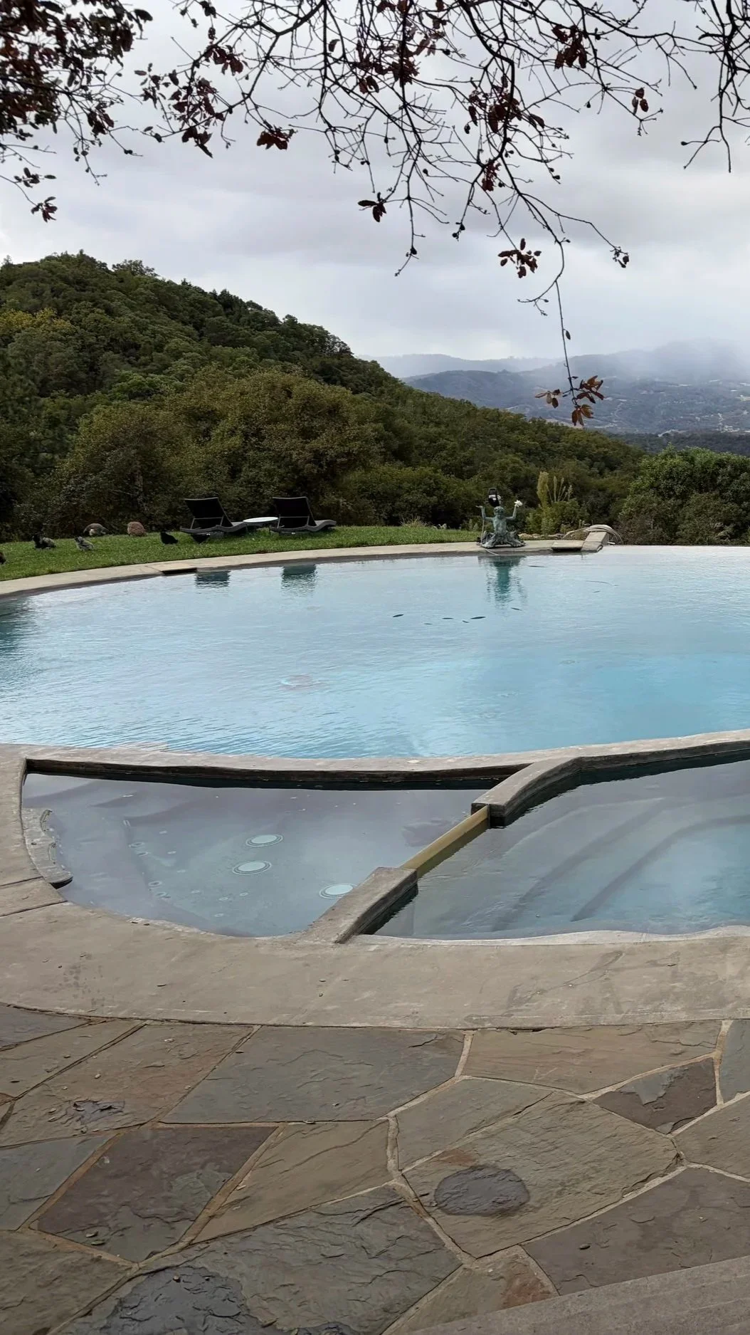 Outdoor pool and hot tub with mountain view, lounge chairs, a swimming pool, and a fountain amidst lush greenery and hilly landscape.