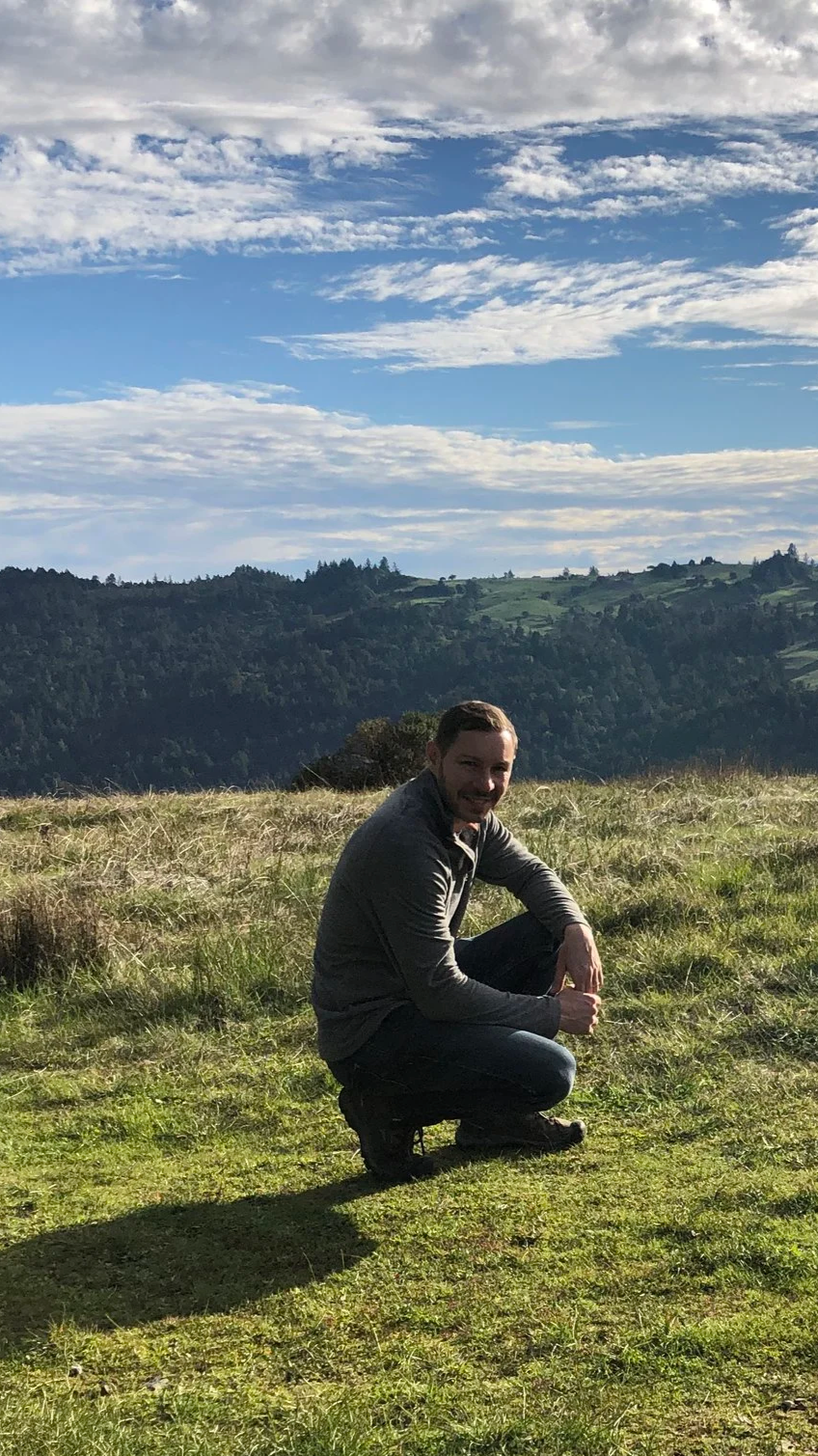 A man kneeling on grass in a scenic outdoor setting at the top of the Islands in the Sky hike, with rolling hills and partly cloudy sky in the background.