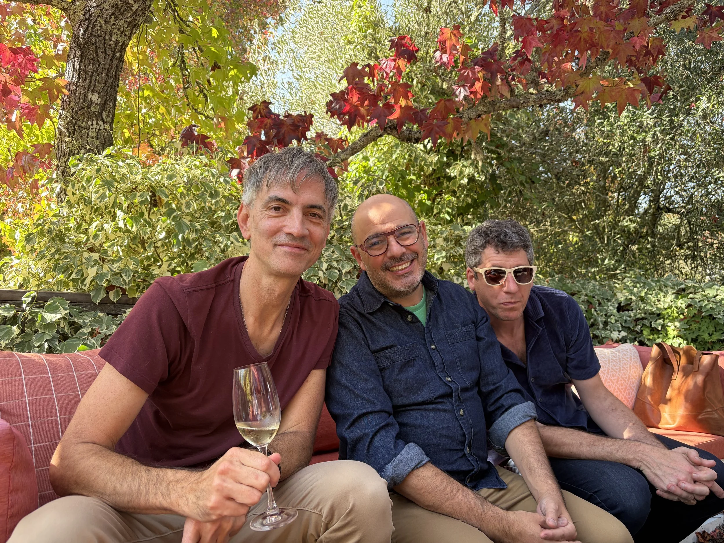 Three men sitting outdoors . The man on the left is smiling and holding a glass of white wine, wearing a maroon shirt. The man in the middle is smiling, wearing glasses and a dark blue jacket. The man on the right is wearing sunglasses and a navy shirt. They are surrounded by lush trees with red and green leaves and sunlight filtering through the branches.