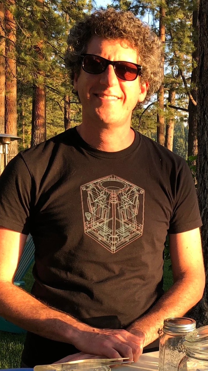 Chef David Karlin wearing sunglasses and a black T-shirt with geometric design smiling outdoors surrounded by trees.