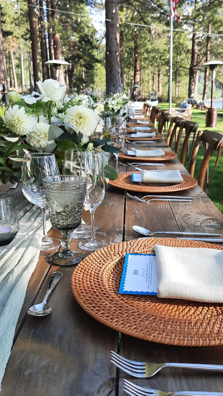 A long outdoor dining table amidst the redwoods set for a gathering, decorated with a floral centerpiece, glassware, wicker placemats, and napkins, surrounded by wooden chairs, with trees and greenery in the background.