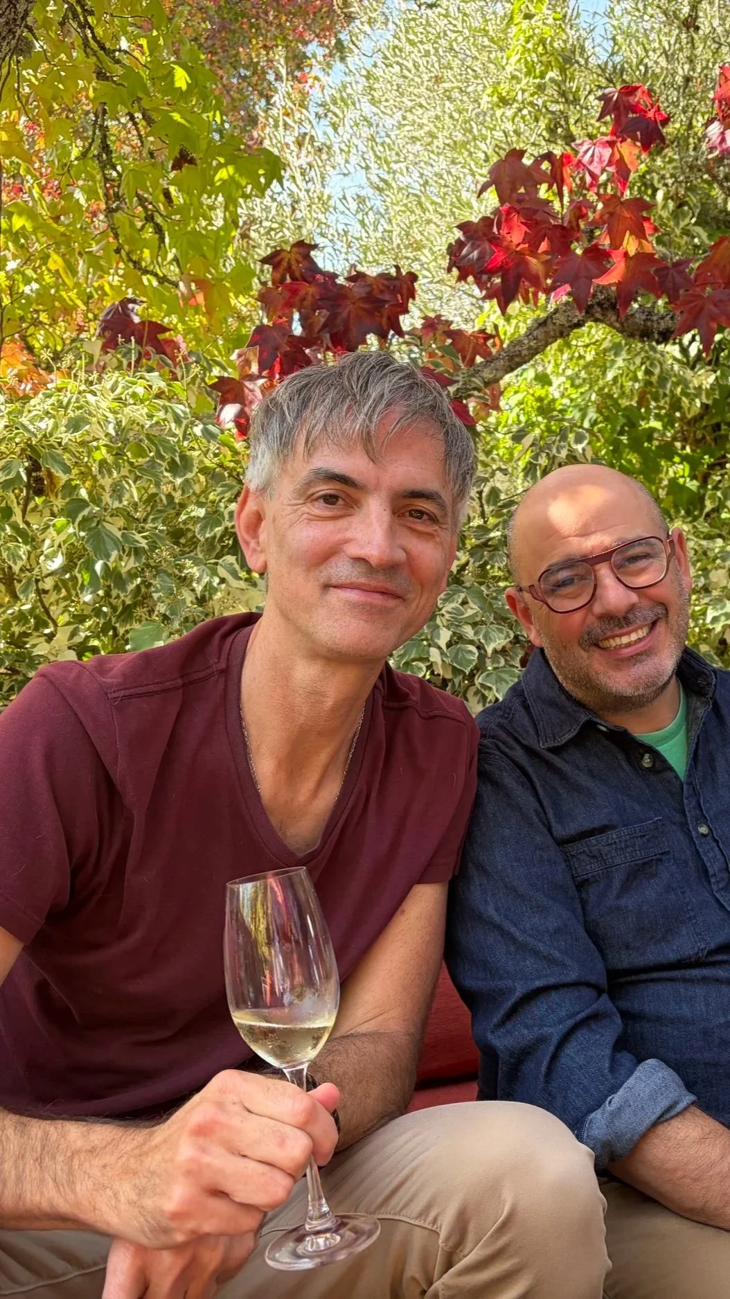 Two men sitting outdoors at Iron Horse Winery among greenery, one holding a glass of white wine, smiling at the camera.