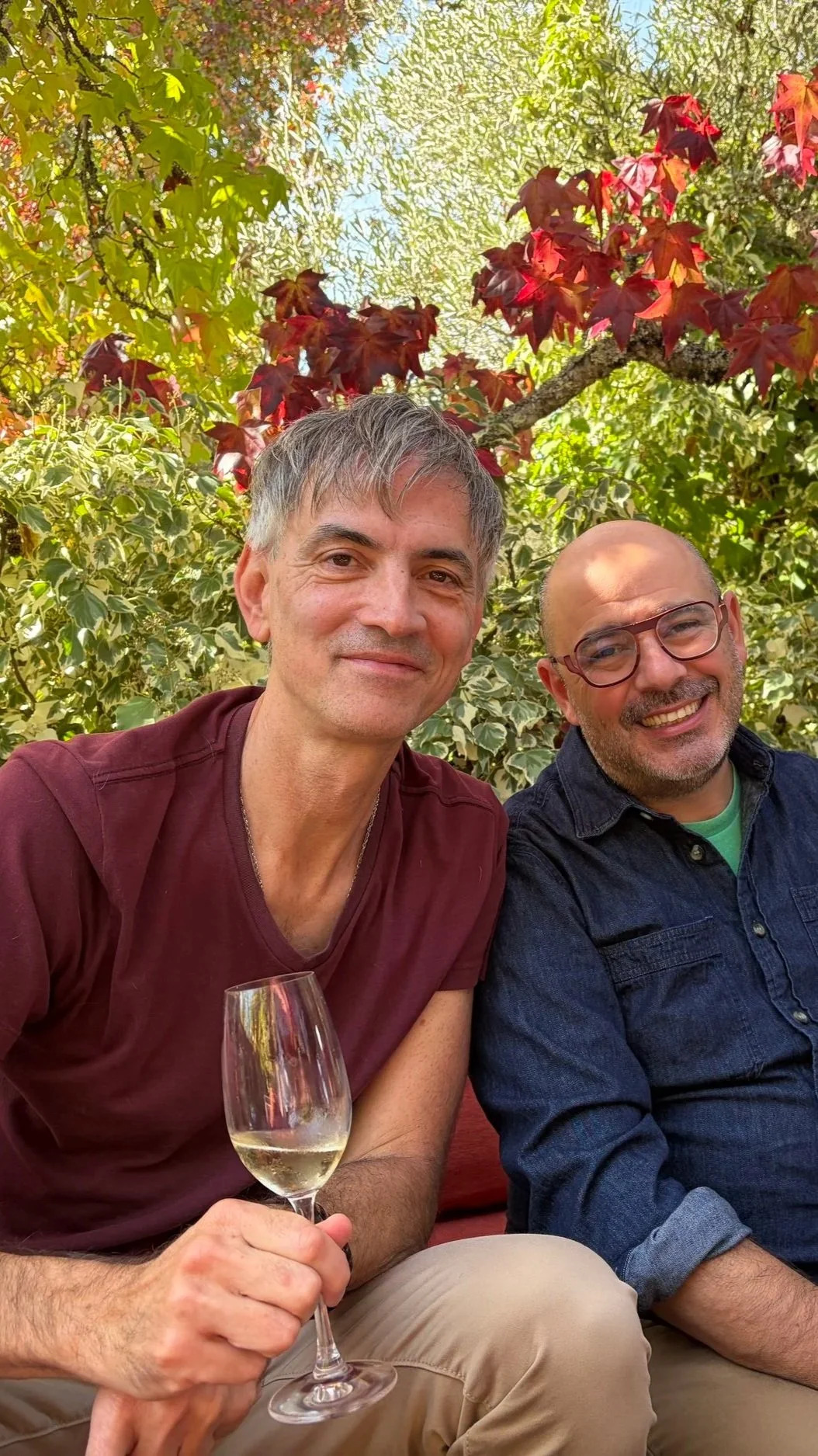 Two middle-aged men sitting outdoors at Iron Horse winery in front of lush green and red foliage, smiling at the camera. The man on the left is holding a glass of white wine.