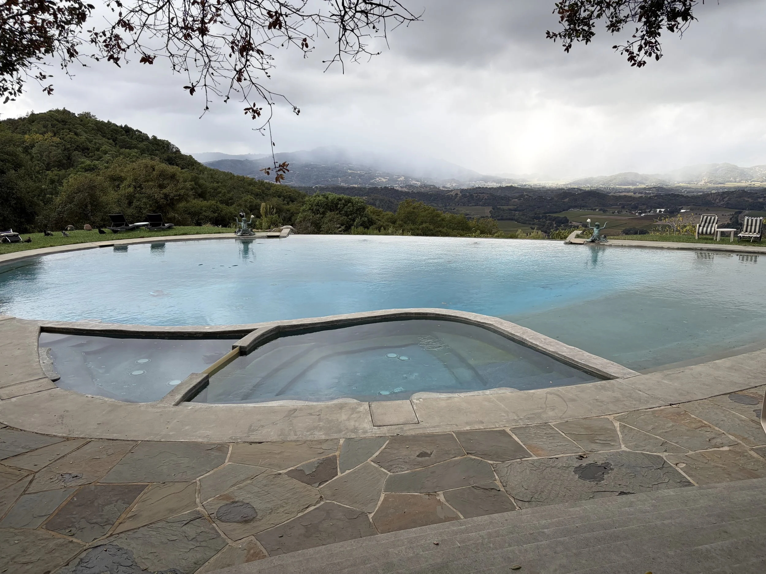 A swimming pool with a view of a hilly landscape and cloudy sky, bordered by stone tiles with outdoor chairs and decorative statues.