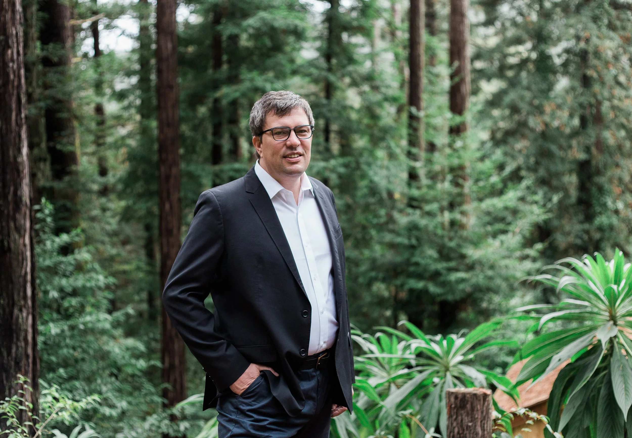 Rob O'Shaughnessy wearing glasses and a black suit, standing outdoors in front of a forest of tall trees and greenery.