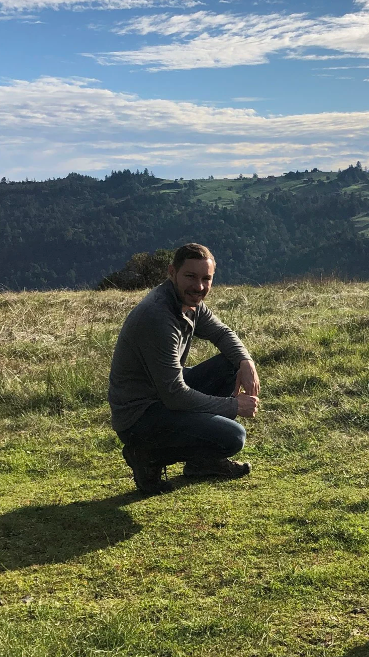 A man squatting on a grassy field with a backdrop of green rolling hills and a partly cloudy sky.