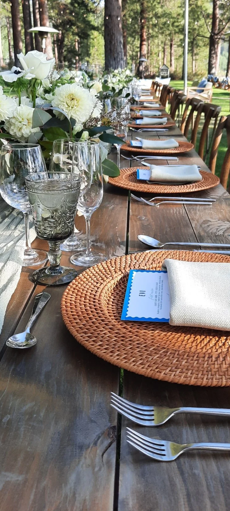 Long outdoor dining table set with floral centerpieces, glassware, plates, napkins, and cutlery, surrounded by wooden chairs in a wooded outdoor setting.
