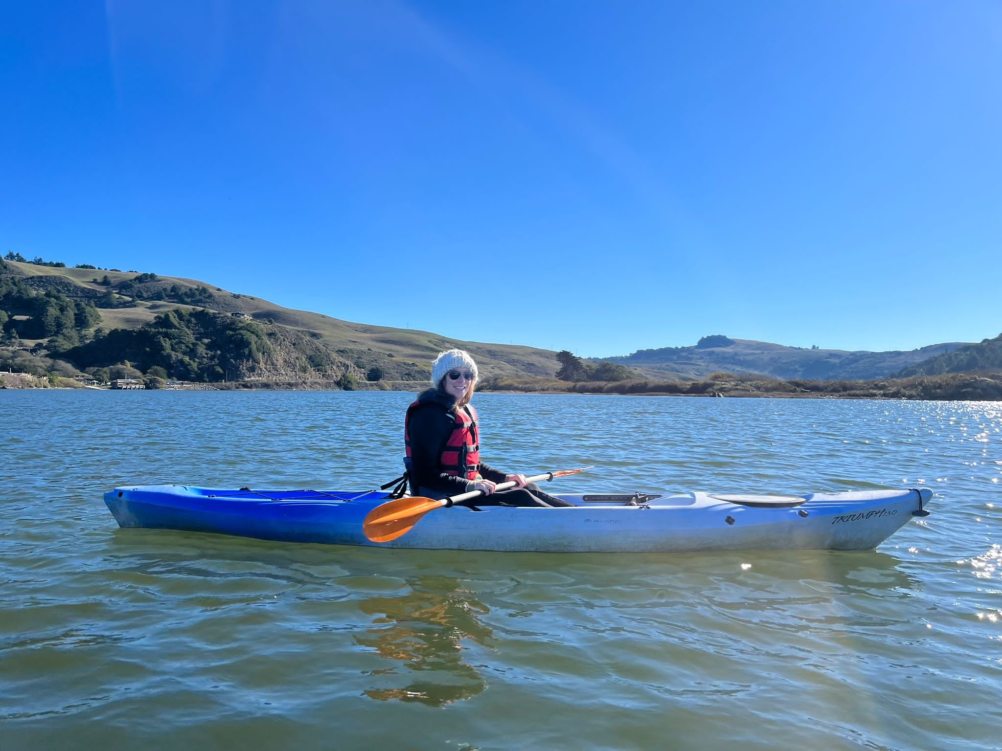 A woman wearing sunglasses, a gray beanie, and a life jacket is sitting in a blue kayak on the calm Russian River. The background features rolling hills with sparse trees and clear blue skies.