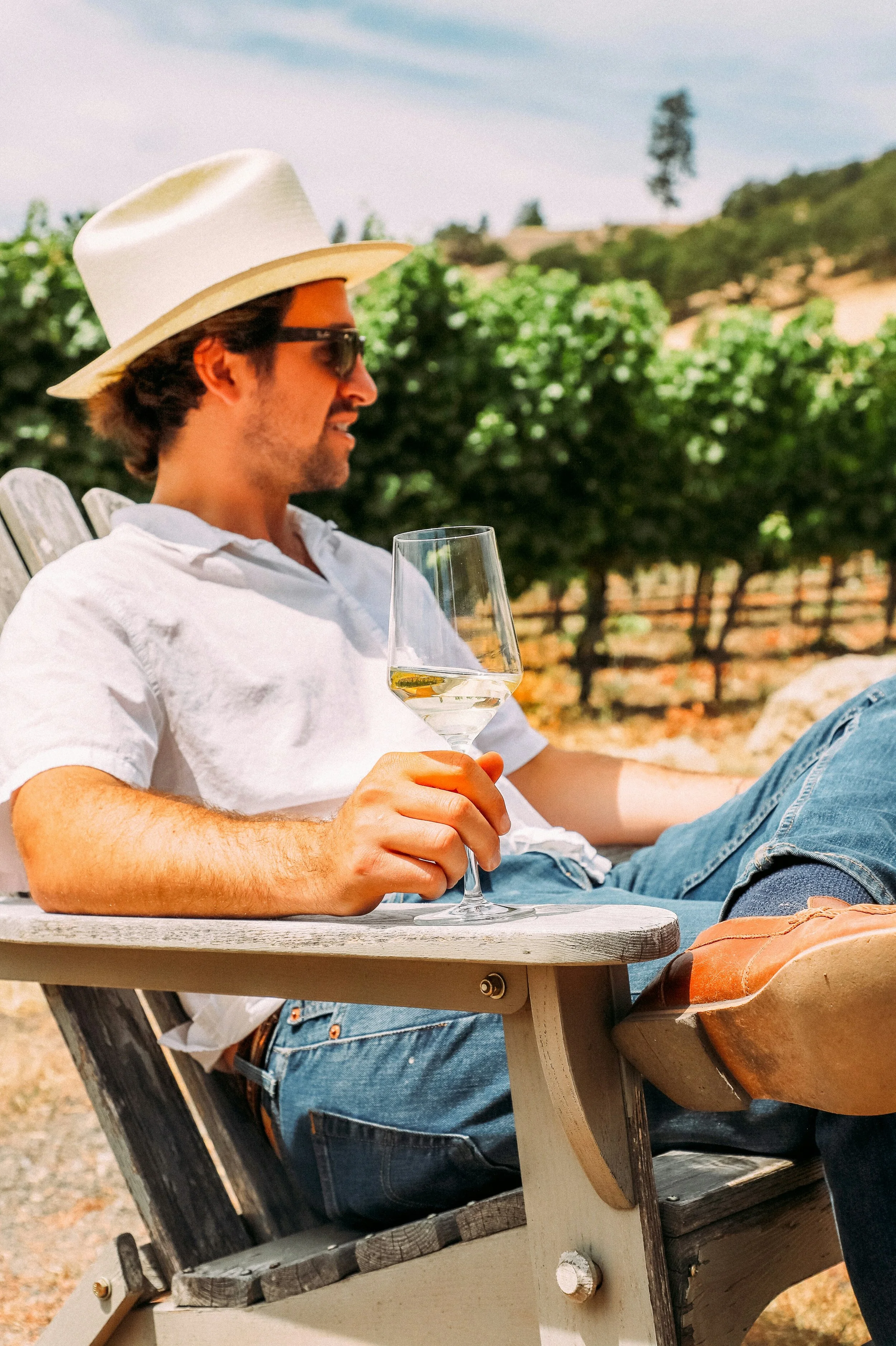 A man wearing a white hat, sunglasses, a white shirt, and jeans relaxing outdoors, holding a glass of white wine, sitting at a wooden picnic table surrounded by greenery and a fence in the background.