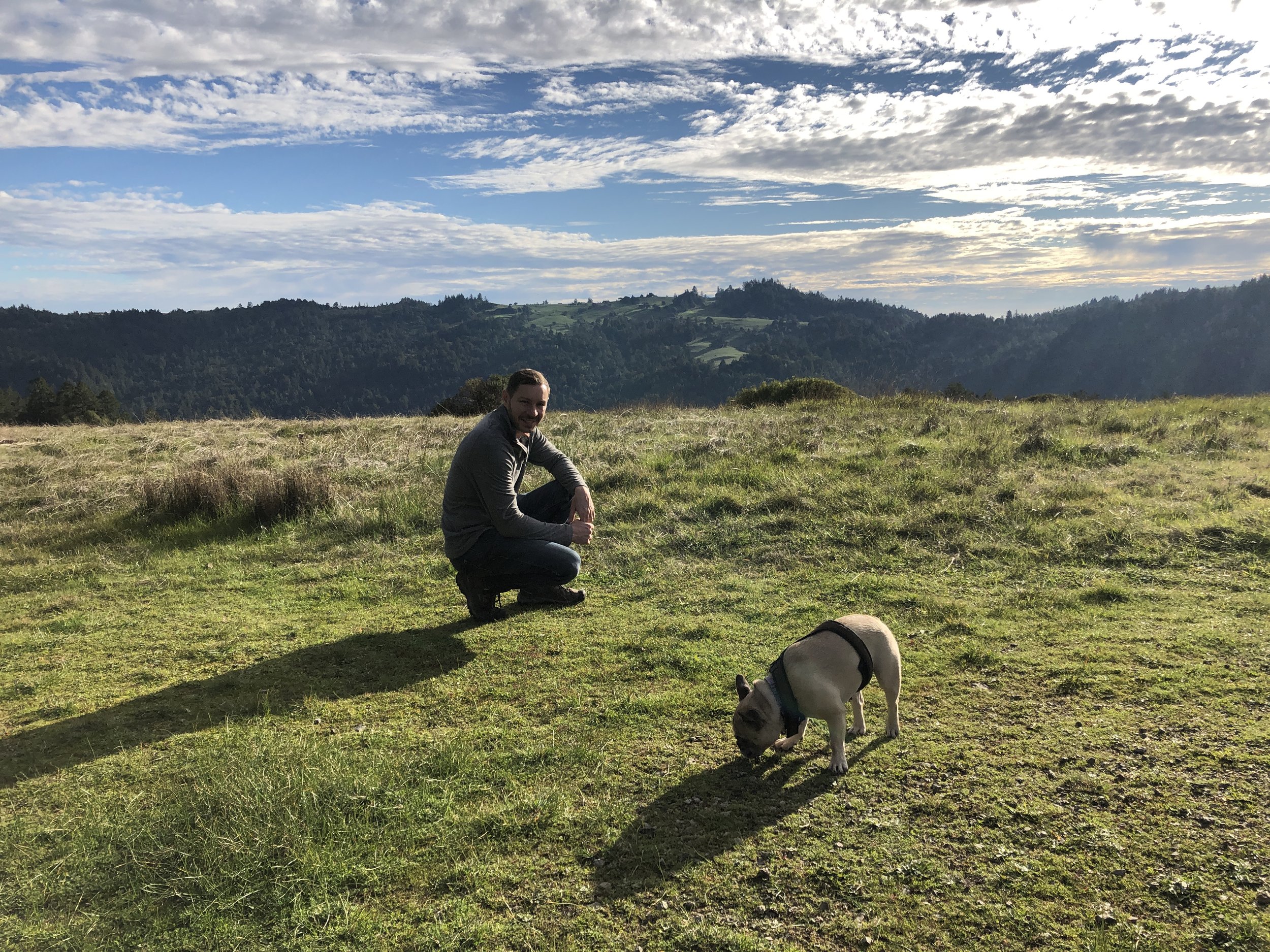 A man sitting on a grassy field with mountains and a partly cloudy sky in the background, alongside a dog wearing a collar.