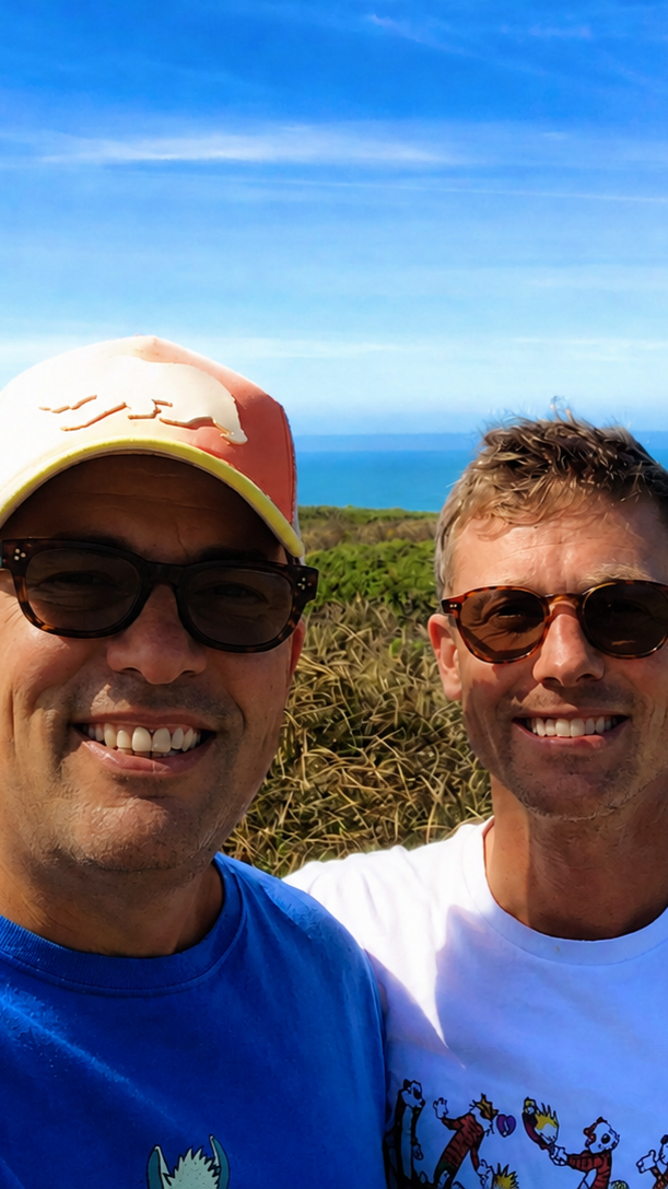 Two men smiling outdoors with a view of the ocean and blue sky in the background. One man is wearing a baseball cap and sunglasses, the other is wearing glasses and sunglasses.