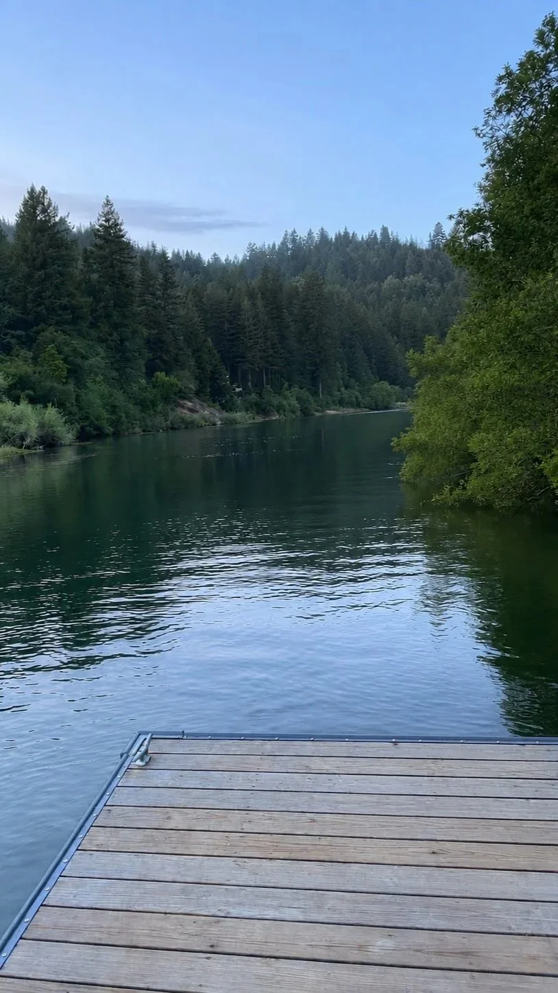 The tranquil Russian River surrounded by dense evergreen trees with a wooden dock extending into the water.