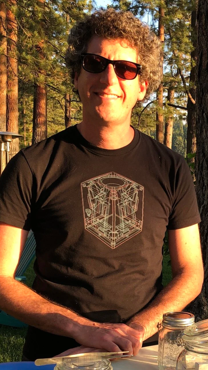 Chef David Karlin hosting a food and wine event wearing sunglasses and a black T-shirt with a geometric design, standing outdoors during sunset in a wooded area with tall pine trees.