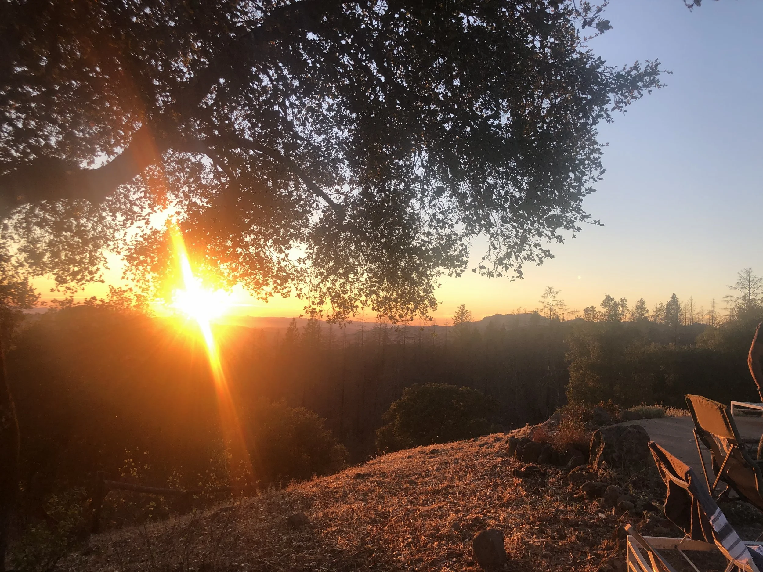 Sunset over a wooded landscape with a picnic chair and a folded camping chair in the foreground.