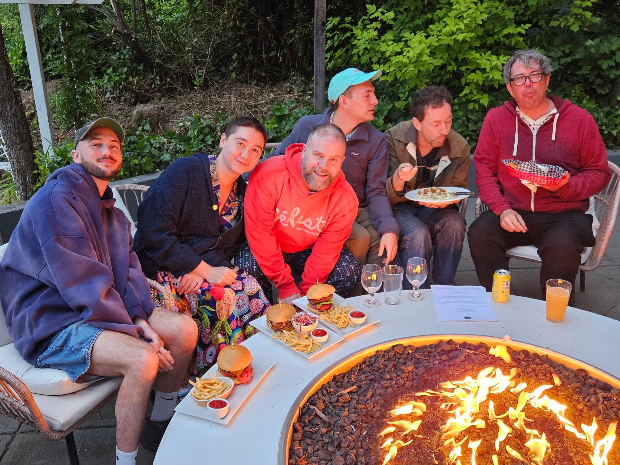 A group of six men sitting around a table outdoors, enjoying food and drinks with a fire pit in the foreground. The table has plates of burgers, fries, and dipping sauces, as well as wine glasses, a can of beer, and a glass of orange juice. The background is filled with green foliage.