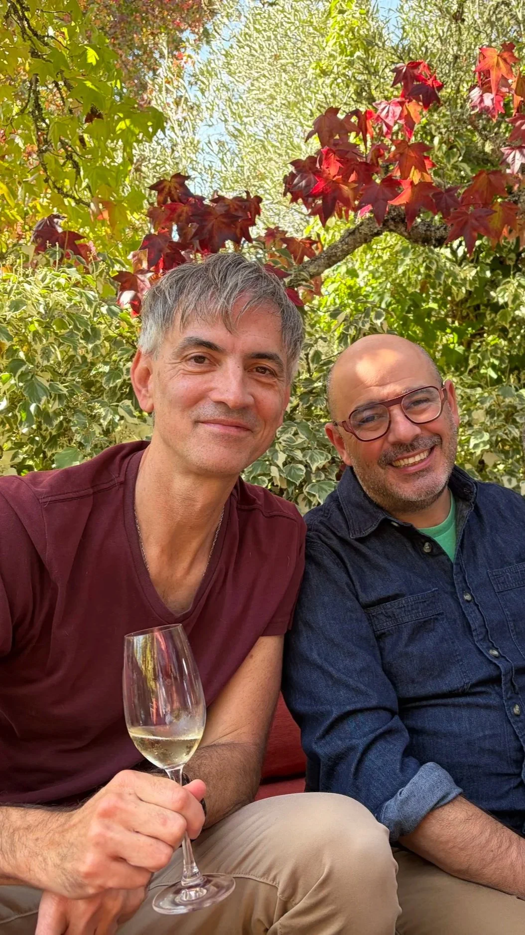 Two men smiling and sitting outdoors tasting wine at Iron Horse Winery among green and red leafy trees, one holding a glass of white wine.
