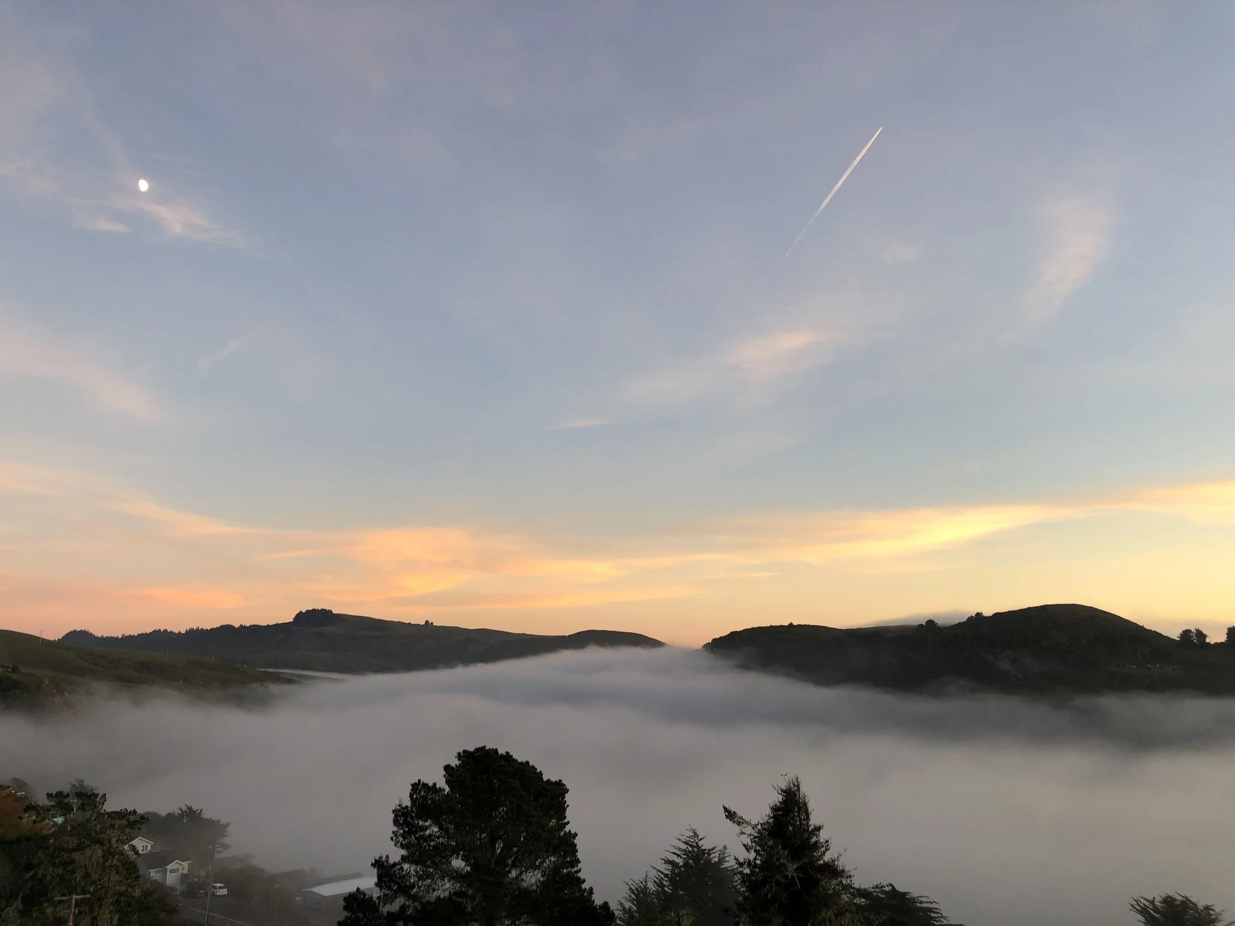 The Russian River shroeded in fog at sunrise with rolling hills, scattered trees, and a few houses in the distance. The sky is partly cloudy with a visible moon and a contrail from an airplane.