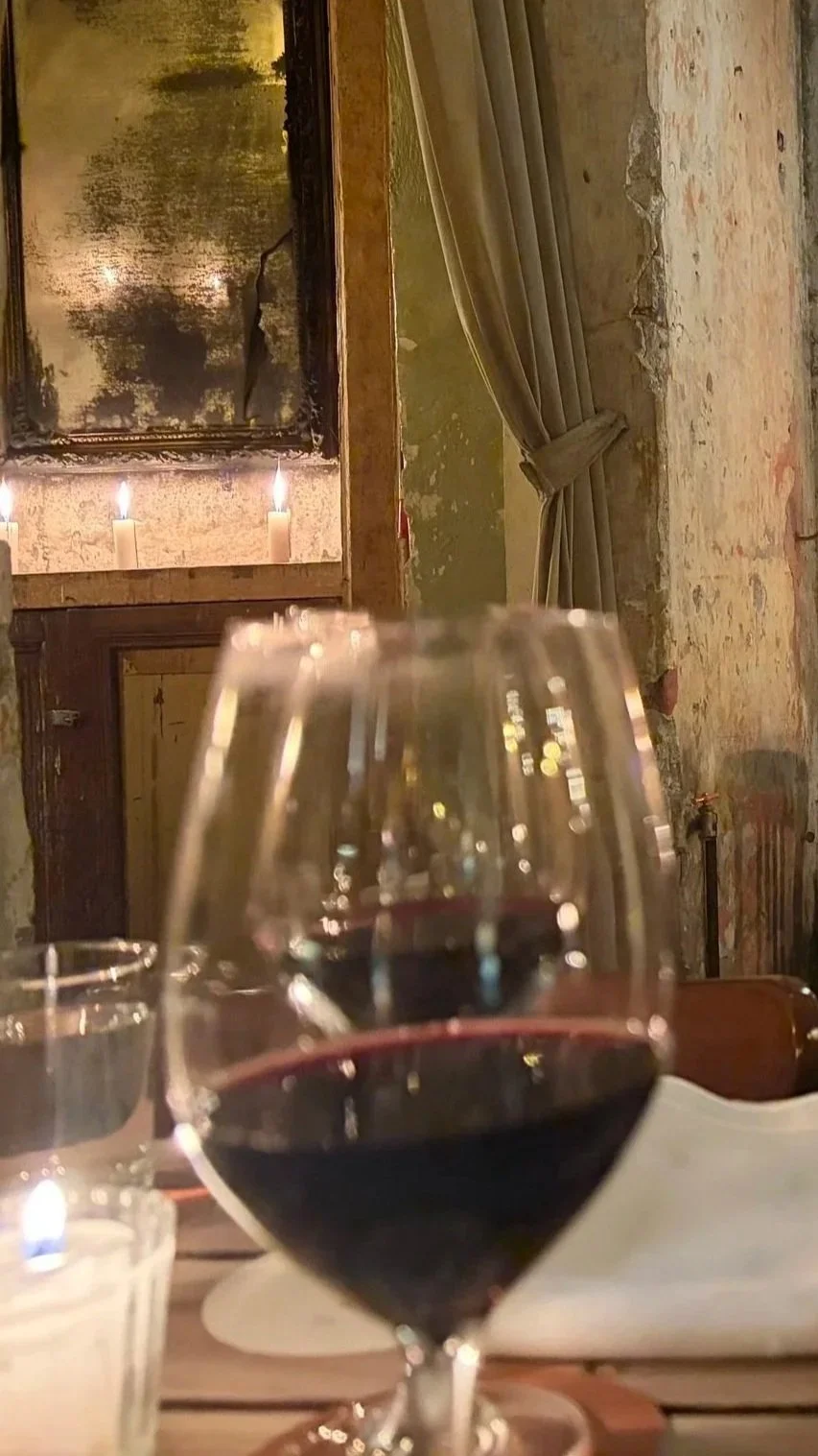 A glass of red wine on a table with candles and a vintage mirror in a rustic, cozy restaurant interior with exposed brick and textured walls.
