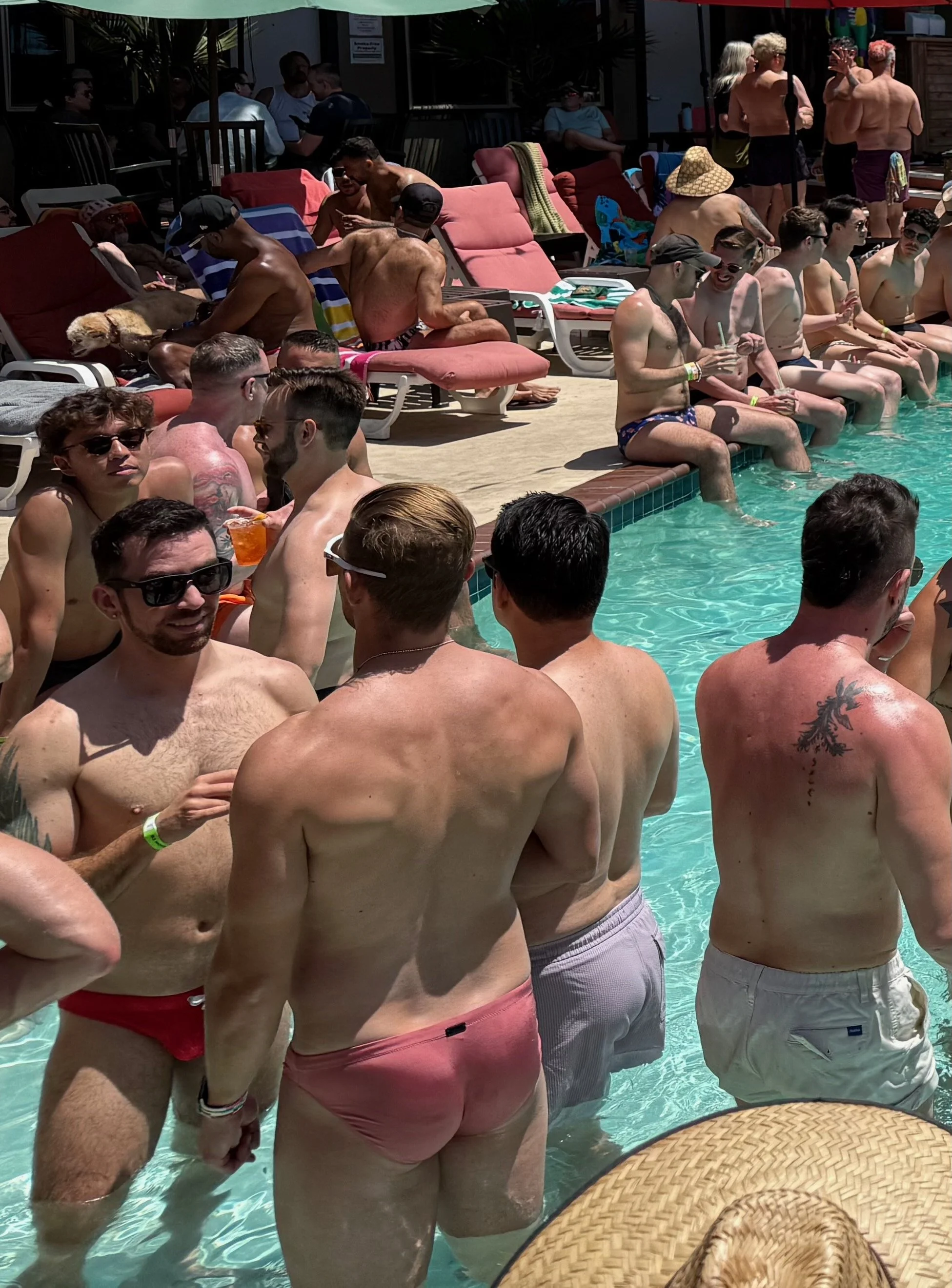People gathered at Guerneville's R3 pool on a sunny day, sitting and standing in shallow water, with some lounging on chairs and others socializing.