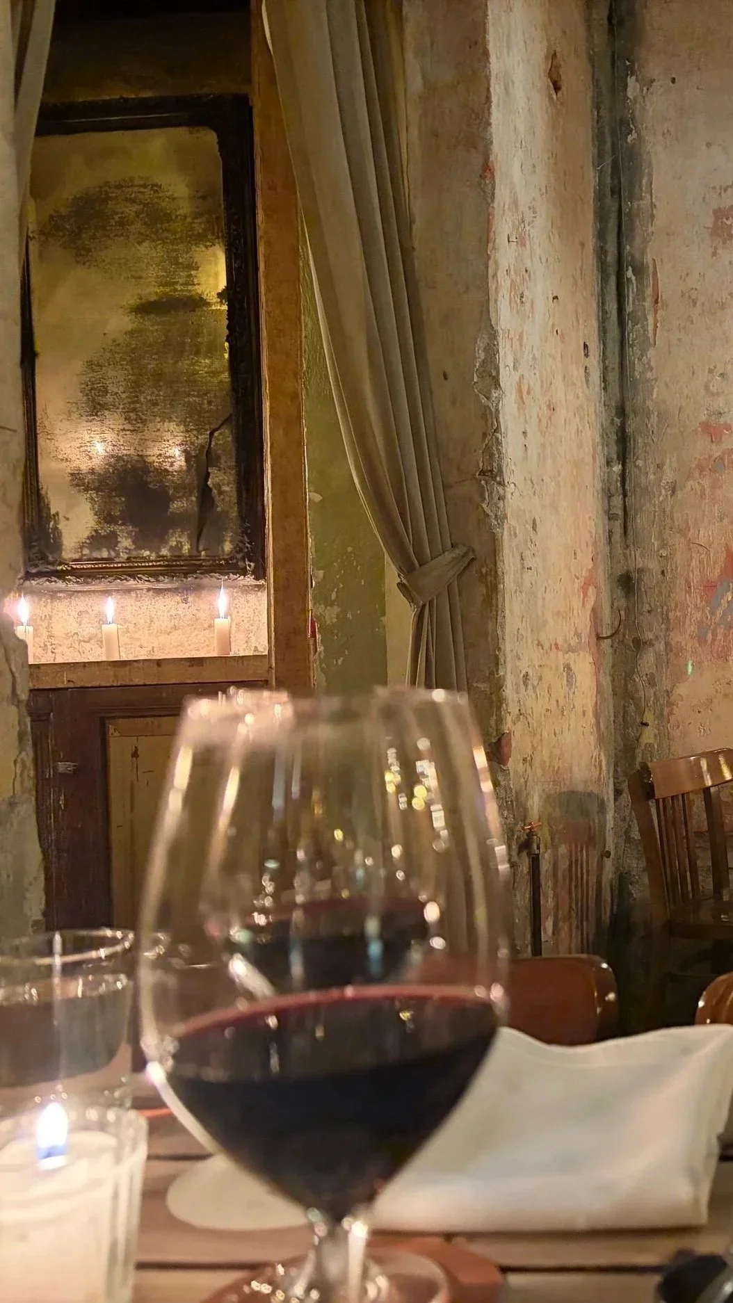 A glass of red wine on a table in a cozy, rustic restaurant with exposed brick walls, a gold-framed mirror, and candles in the background.