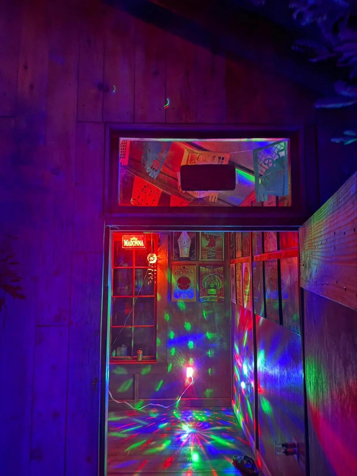 A secret bar with disco lights, a neon sign that says 'Madonna,' posters on the walls, and a mirror reflecting the vibrant lights.