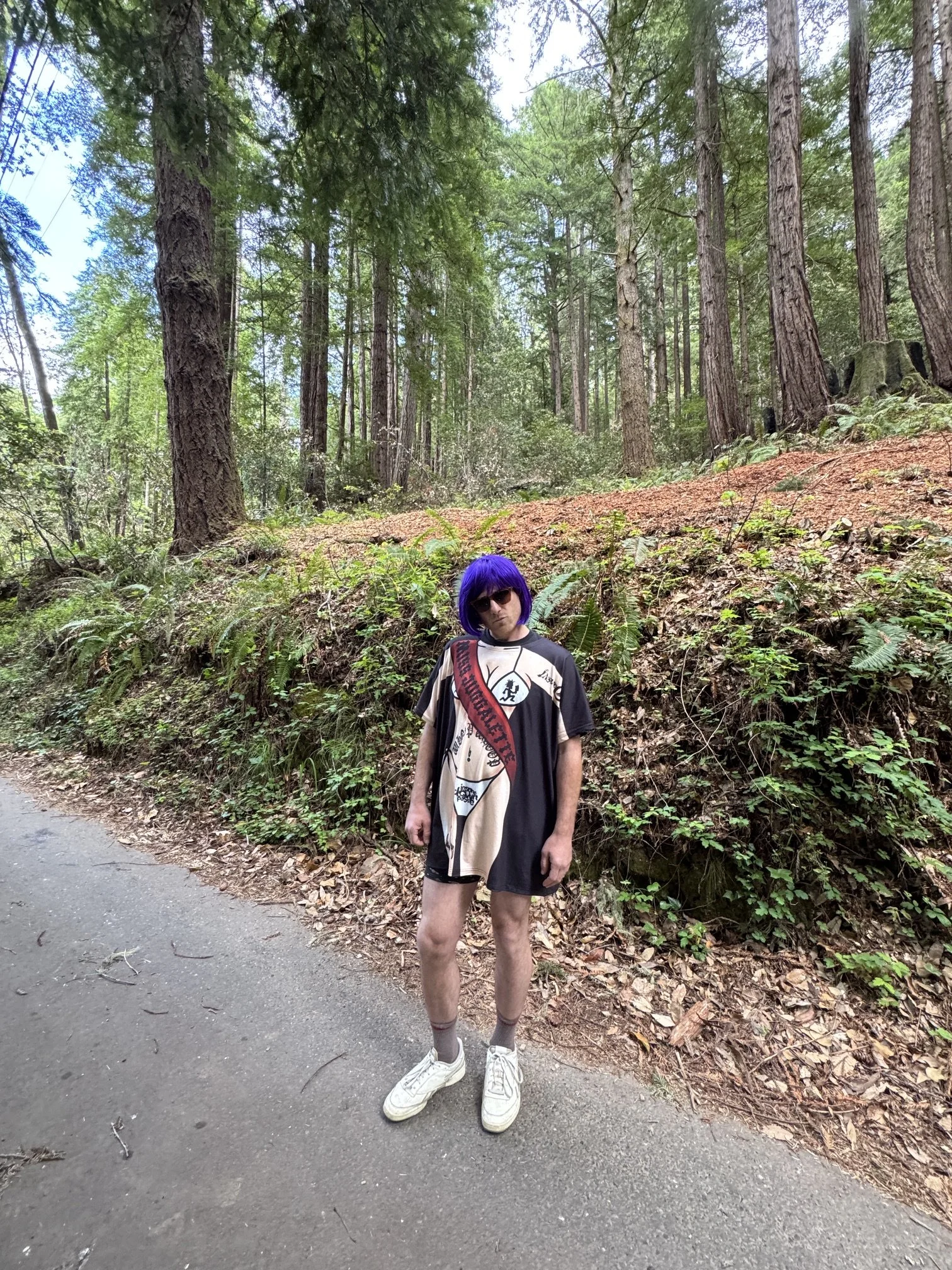 Person with a purple wig, sunglasses, and a graphic T-shirt standing on a paved path in a forest with tall trees and dense greenery.