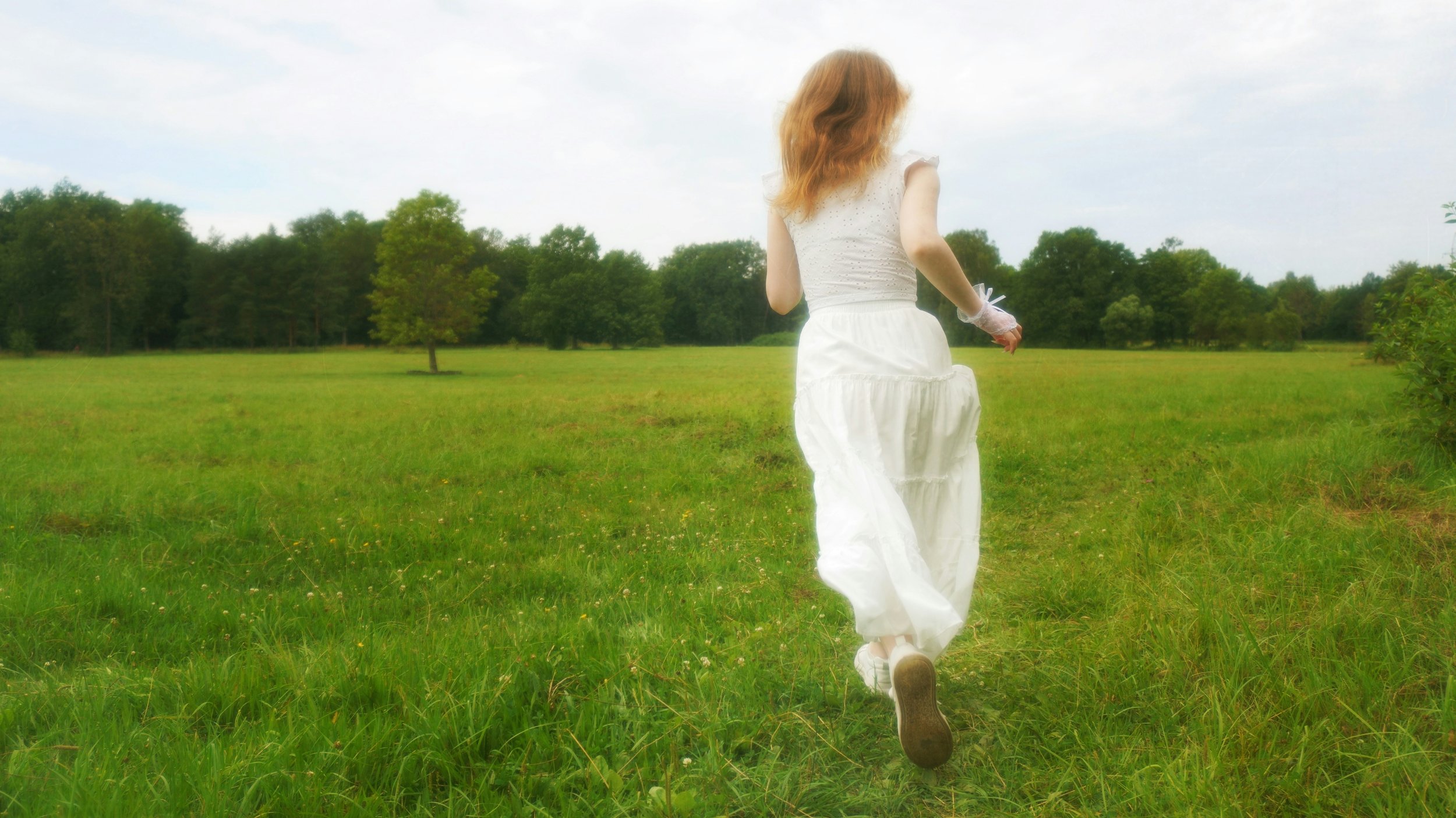 A girl with red hair running through a grassy field wearing a white dress.