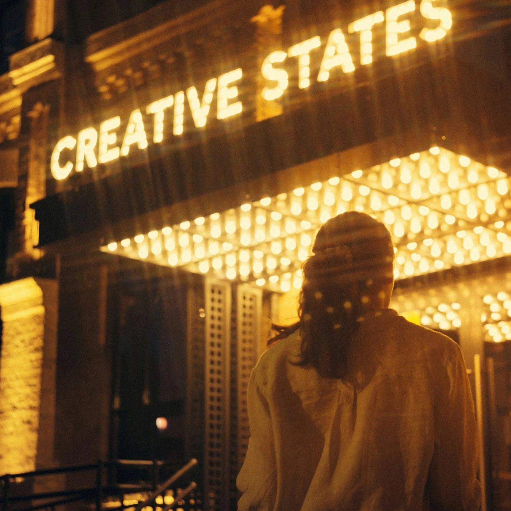 A woman with dark hair and a light-colored jacket walking past a brightly lit sign that says "Creative States" at night, with a canopy of lights overhead.
